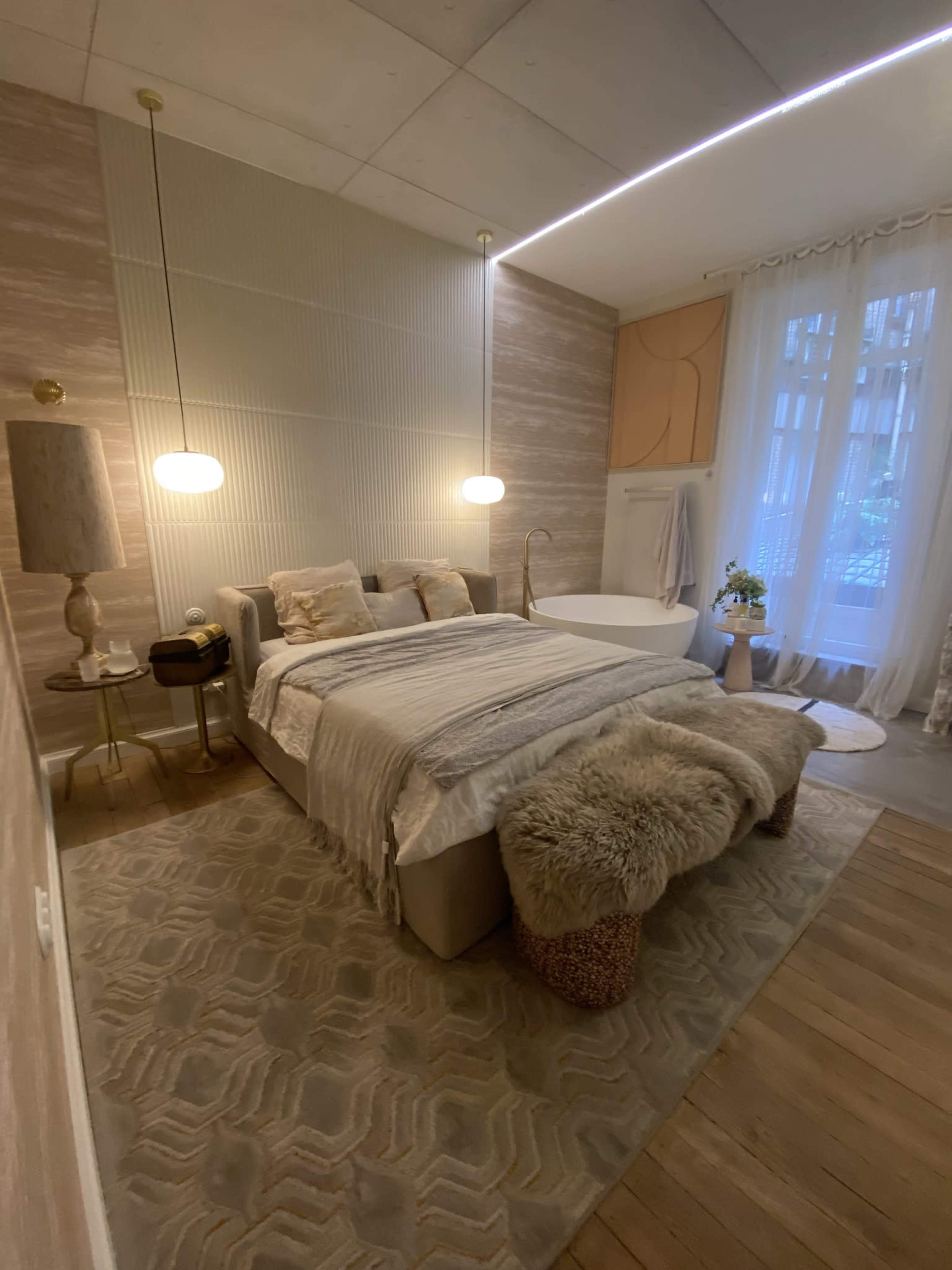 The image shows a modern bedroom featuring a bed with light bedding, a freestanding bathtub, a soft area rug, and large windows allowing natural light to enter.