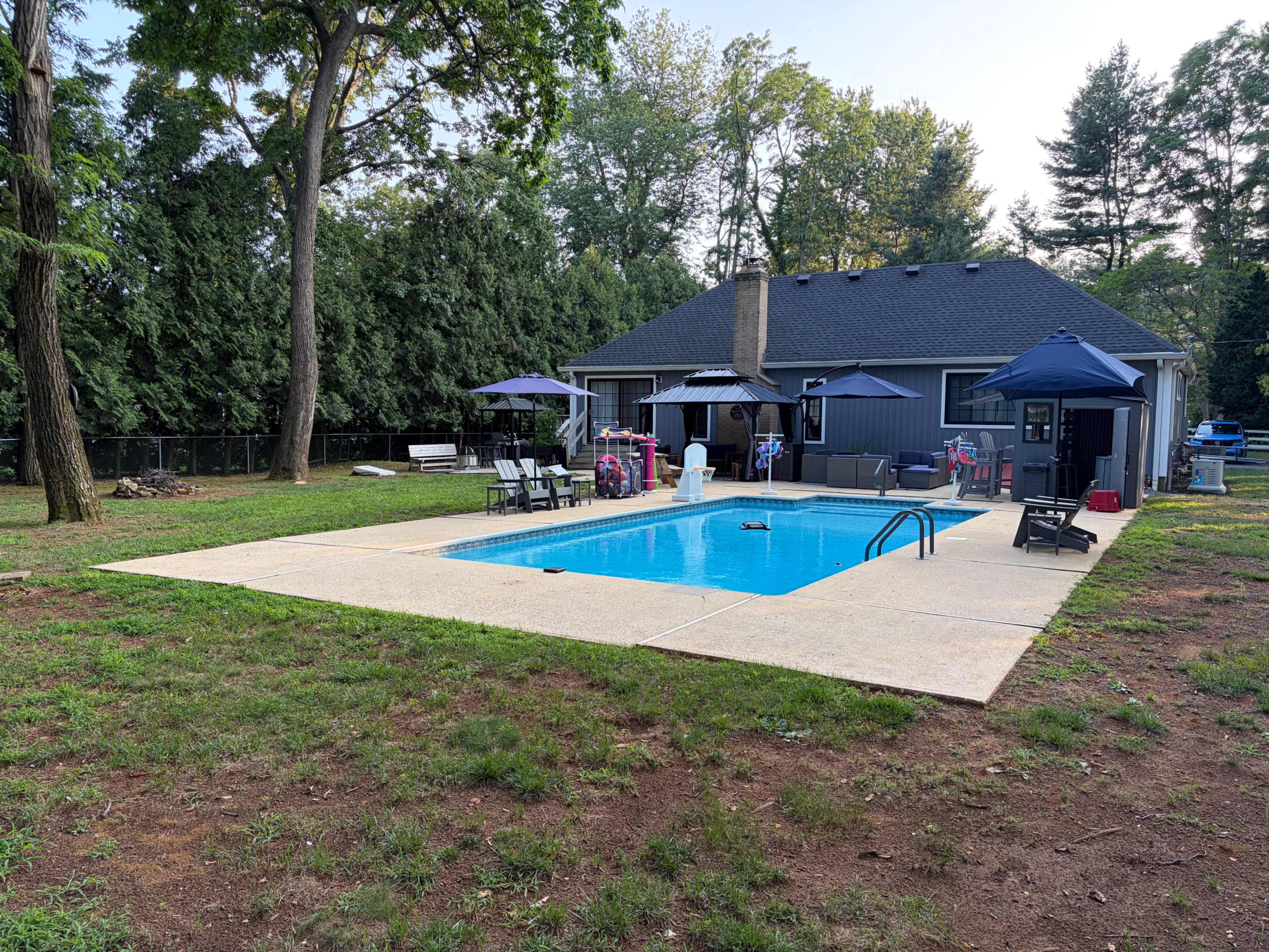 The image shows a backyard with a rectangular swimming pool surrounded by lounge chairs and umbrellas, adjacent to a single-story house.