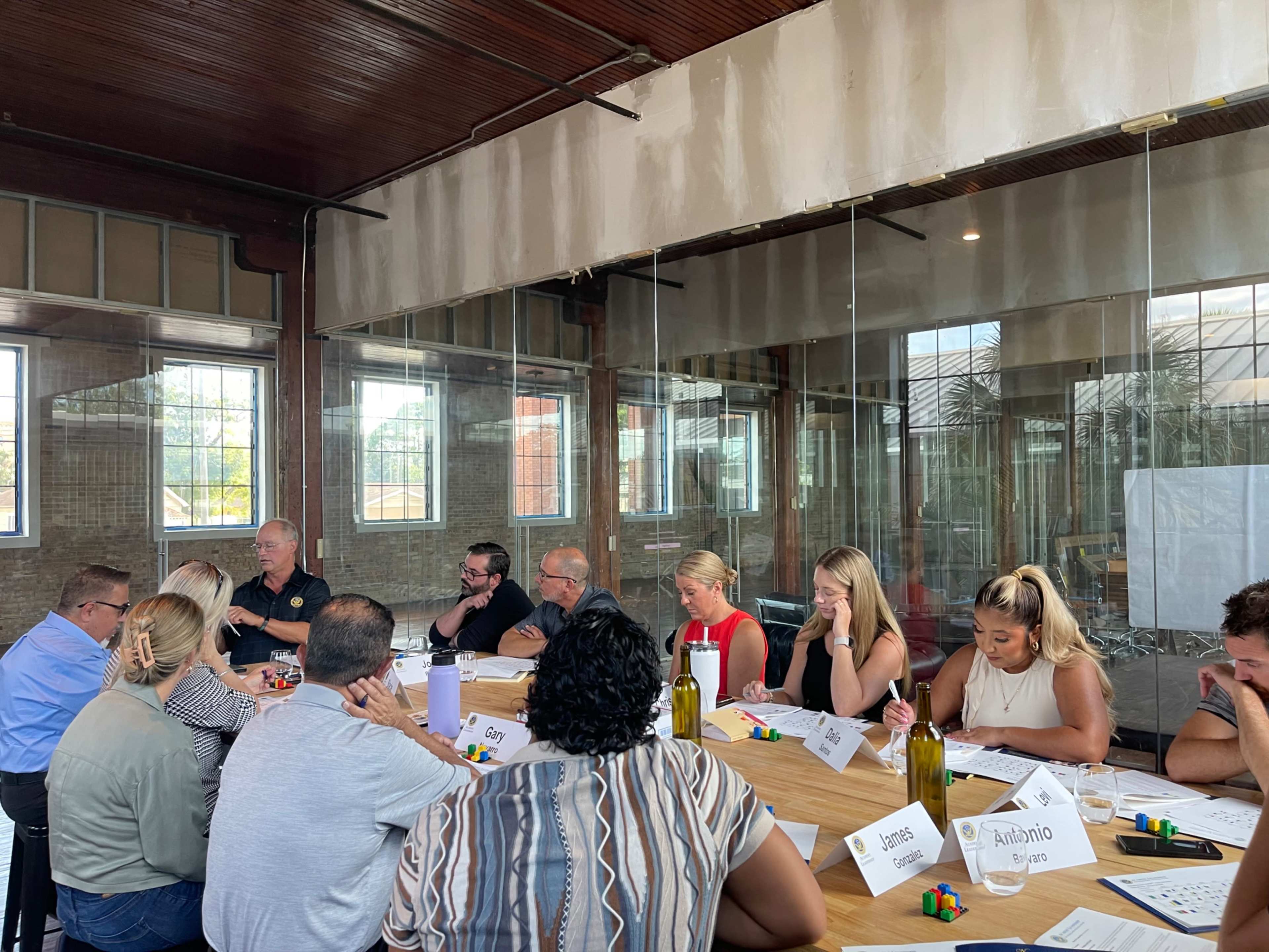 A group of people is seated around a long table in a glass-walled meeting room, engaged in a discussion and taking notes.