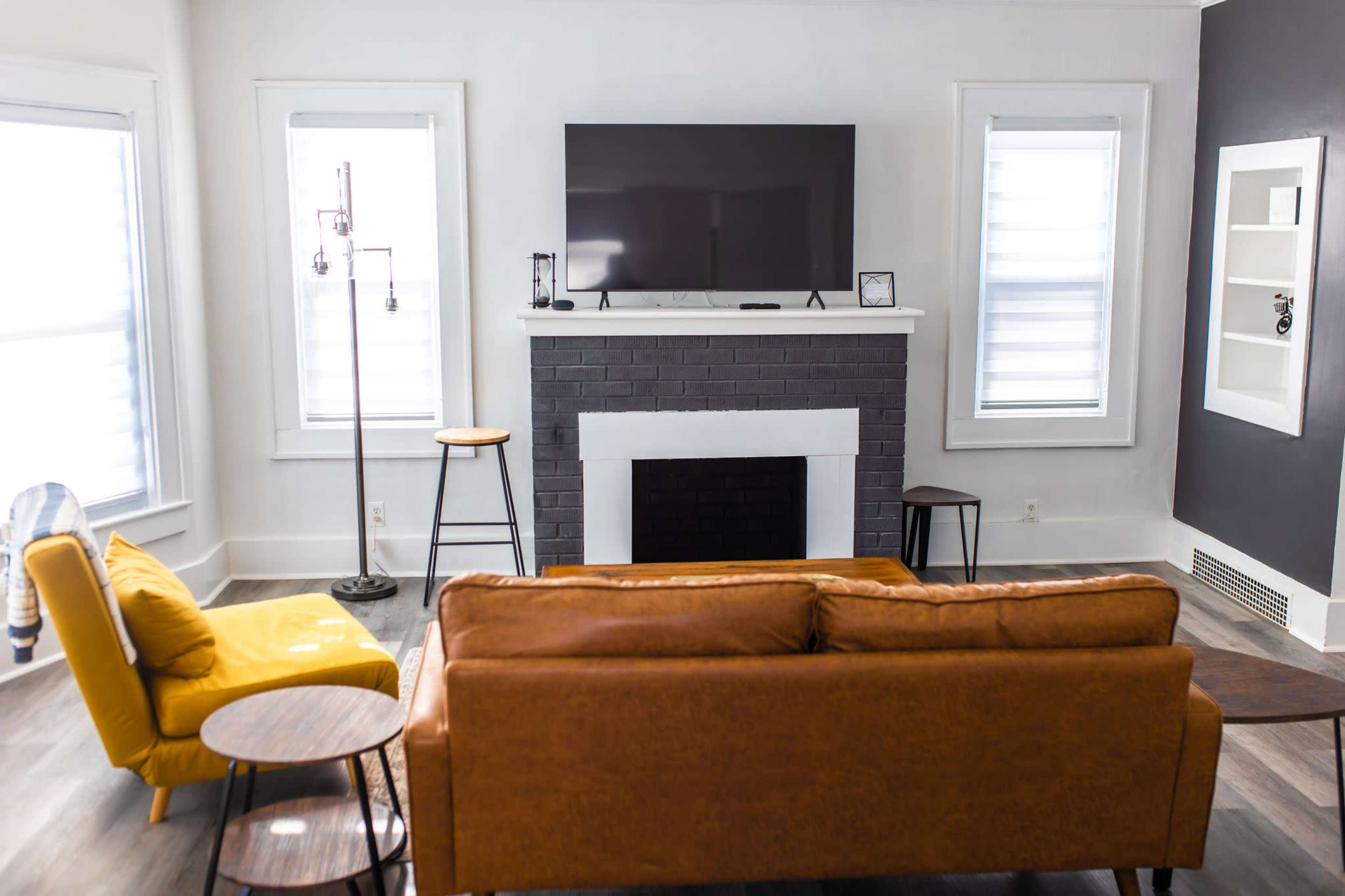 The living room features a brown leather sofa, a yellow armchair, a flat-screen TV mounted above a black brick fireplace, and several wooden stools arranged around the space.