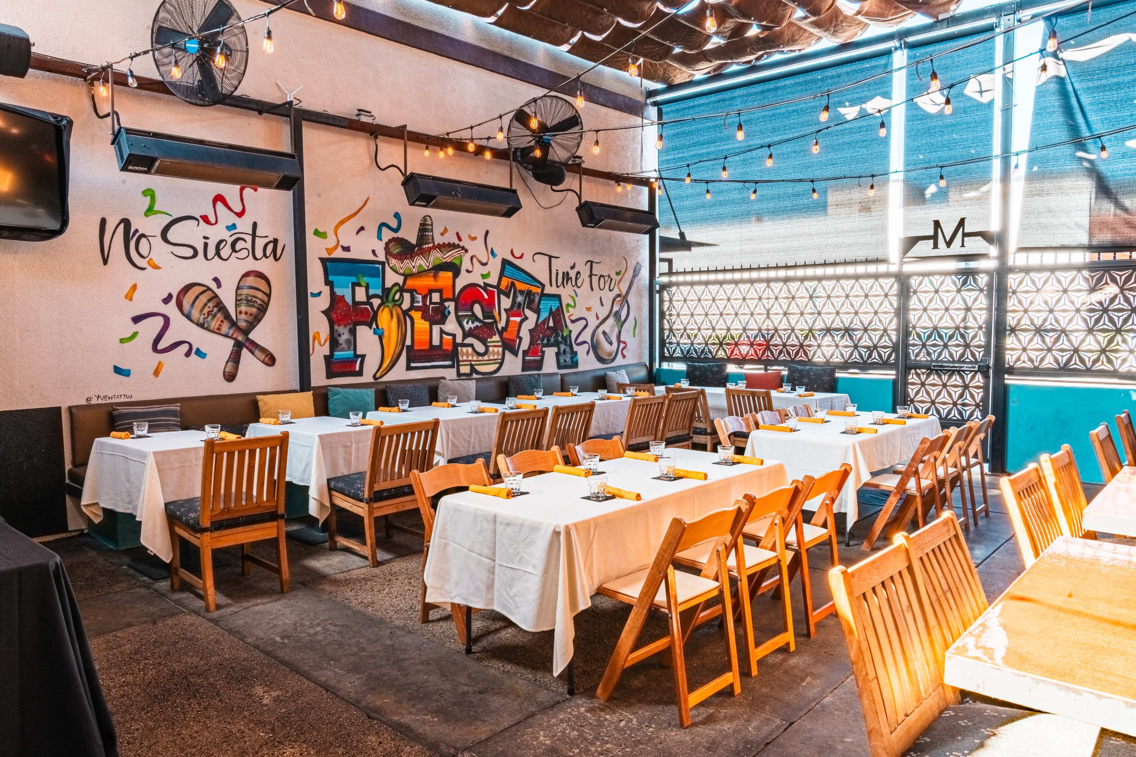 The image shows a brightly lit dining area with multiple tables covered in white linens, featuring colorful murals on the wall and string lights overhead.