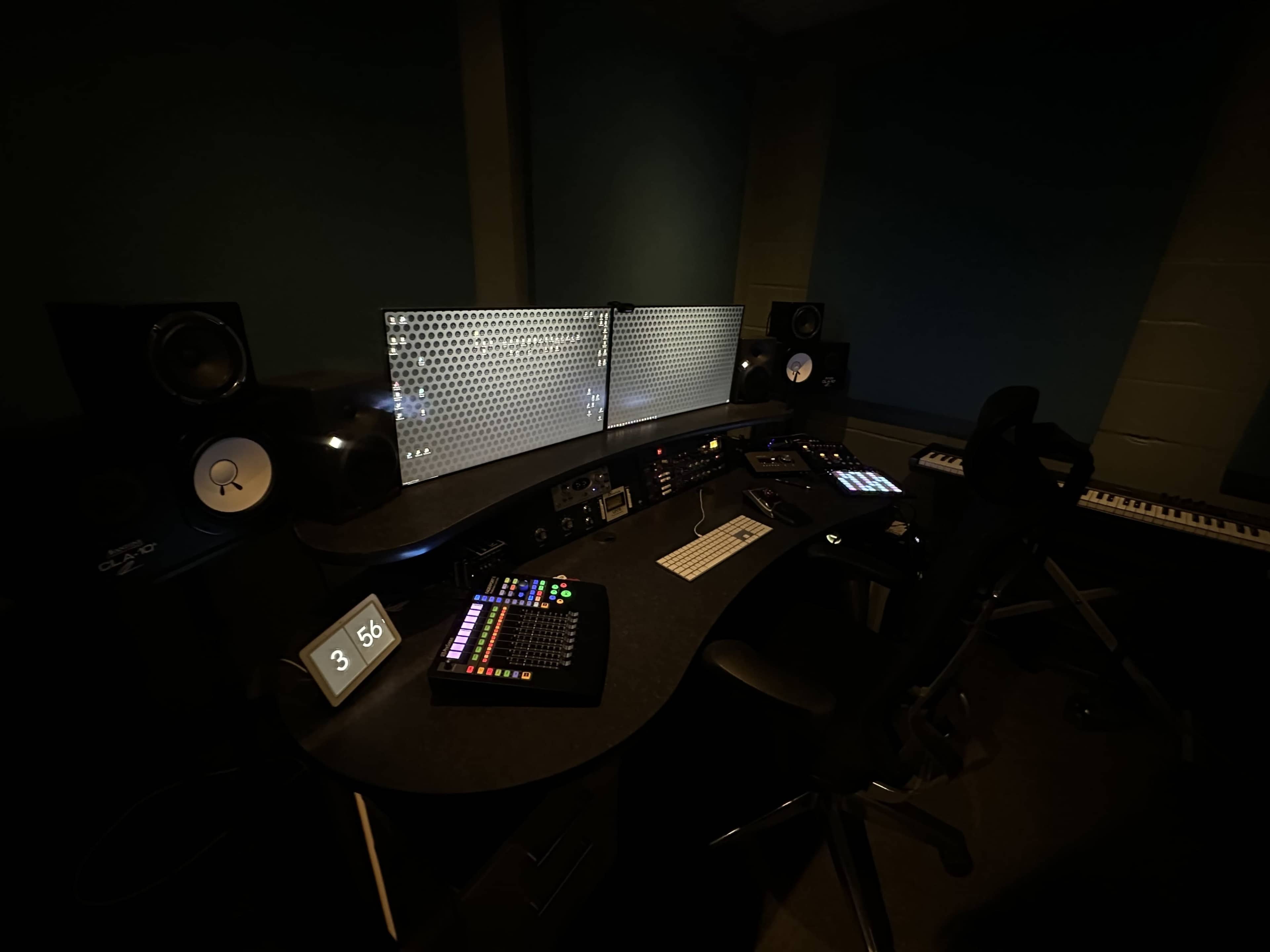 A dimly lit music production studio featuring a mixing console, monitors, a keyboard, and a digital clock displaying the time.