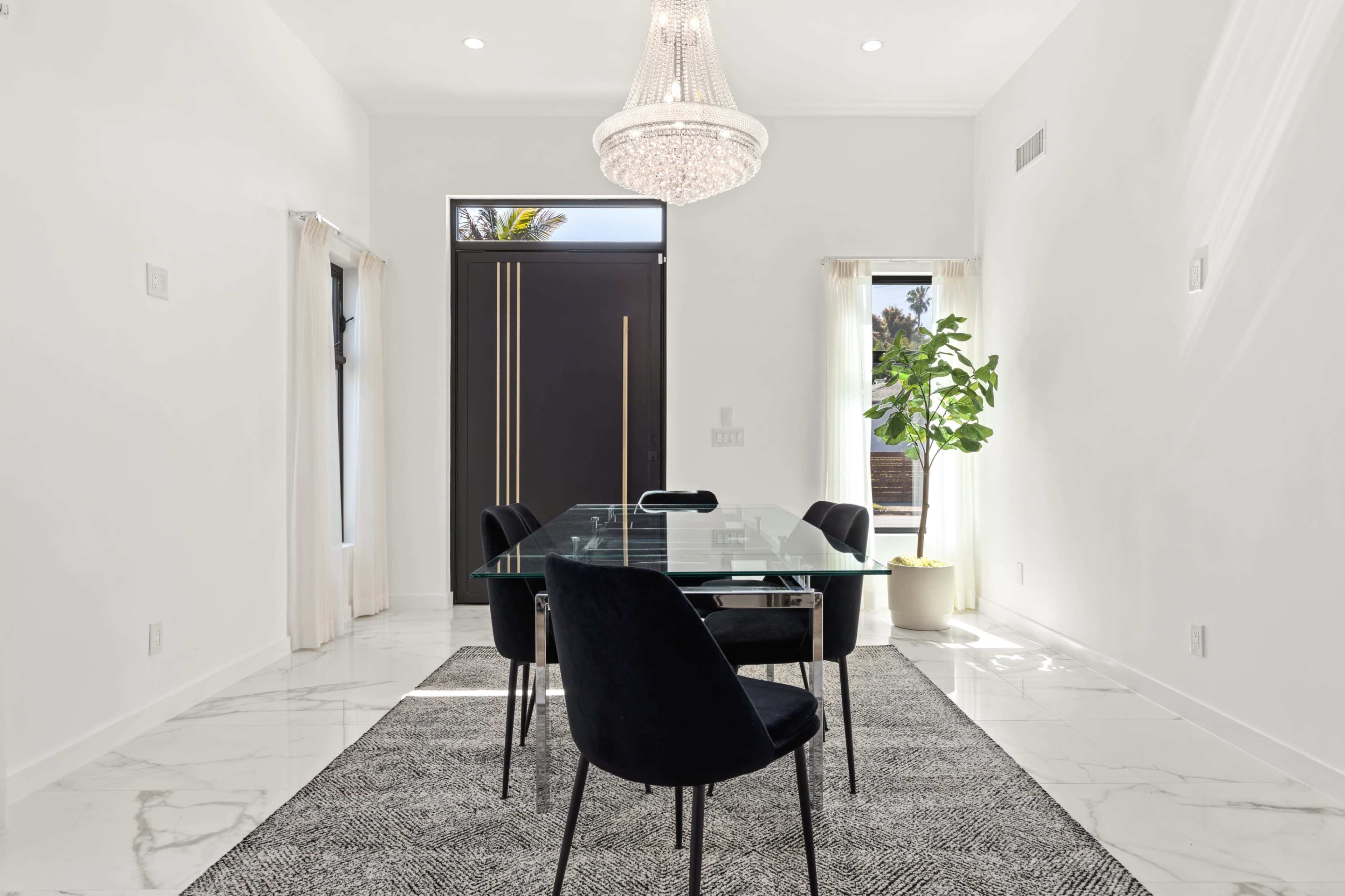 A modern dining room features a glass table surrounded by black chairs, a large rug, and a chandelier, illuminated by natural light from the windows.