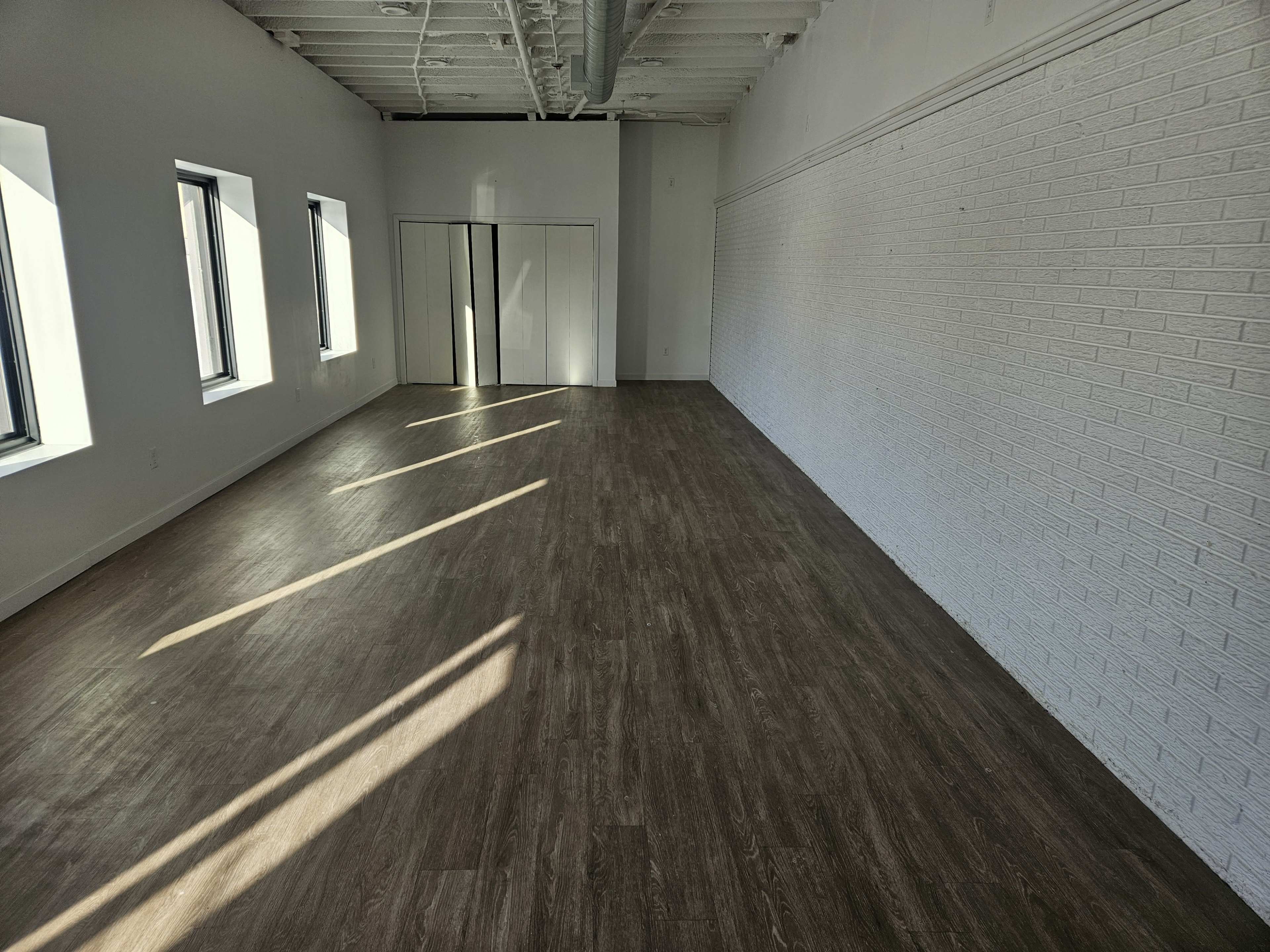 The image shows an empty room with wooden floors, white walls, and large windows allowing sunlight to stream in.