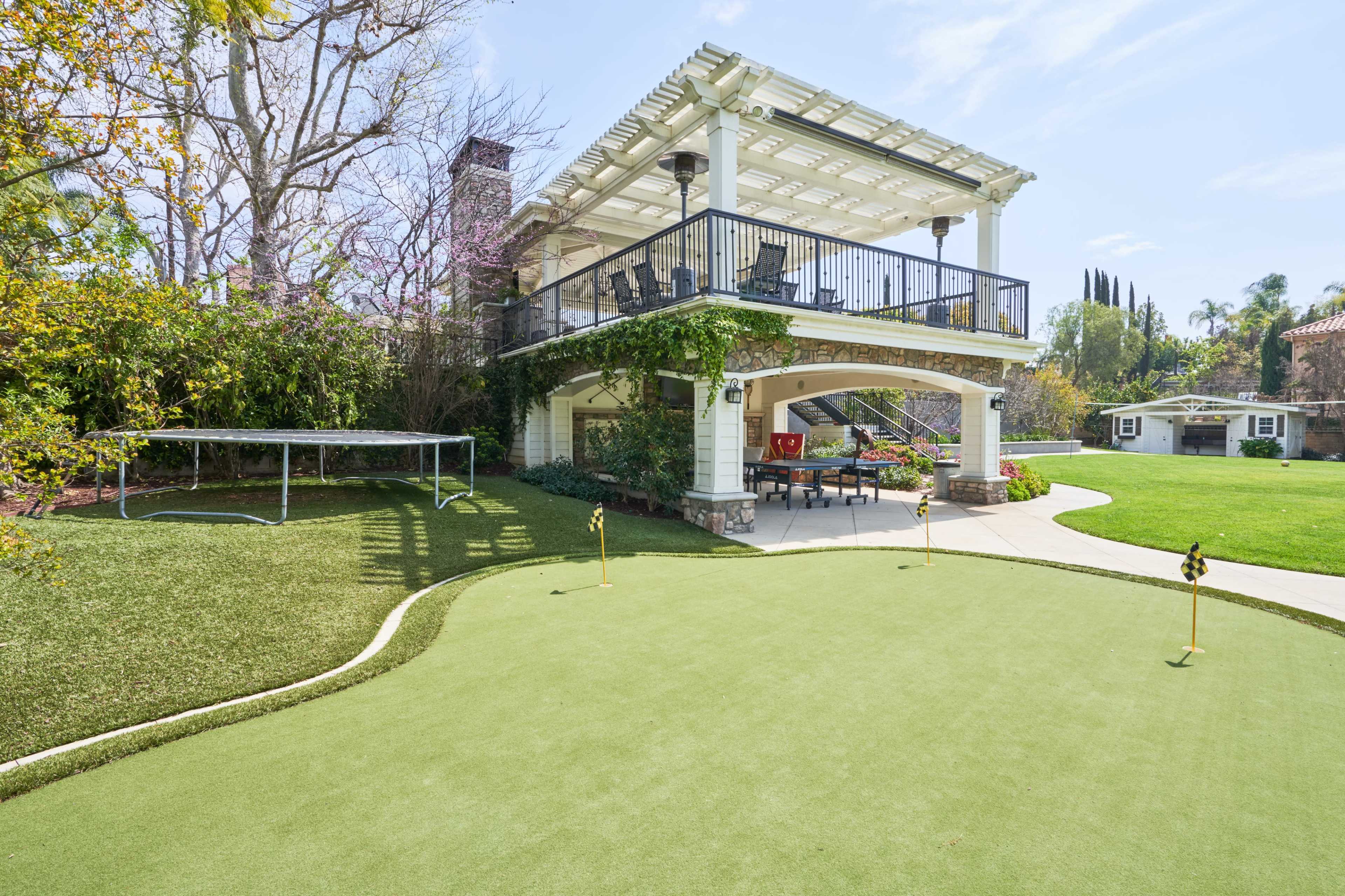 A backyard with a covered patio, a putting green, and a small soccer goal, surrounded by well-maintained landscaping.
