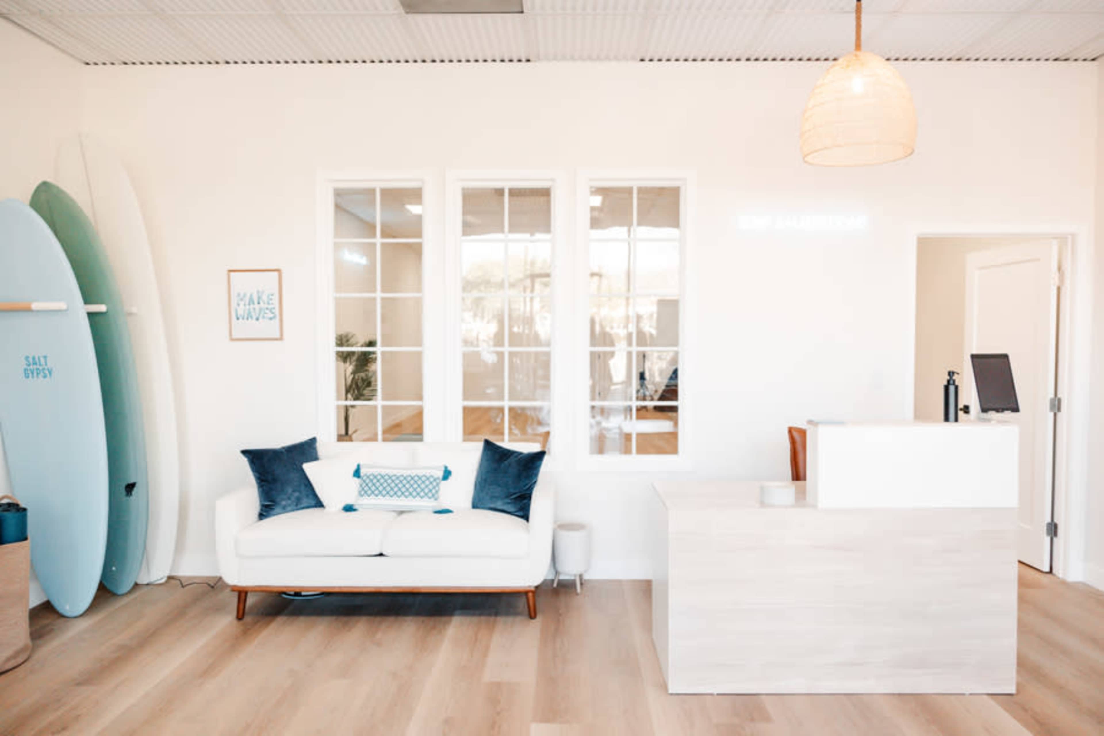 The image shows a bright reception area with a sofa, surfboards on the wall, and a reception desk.
