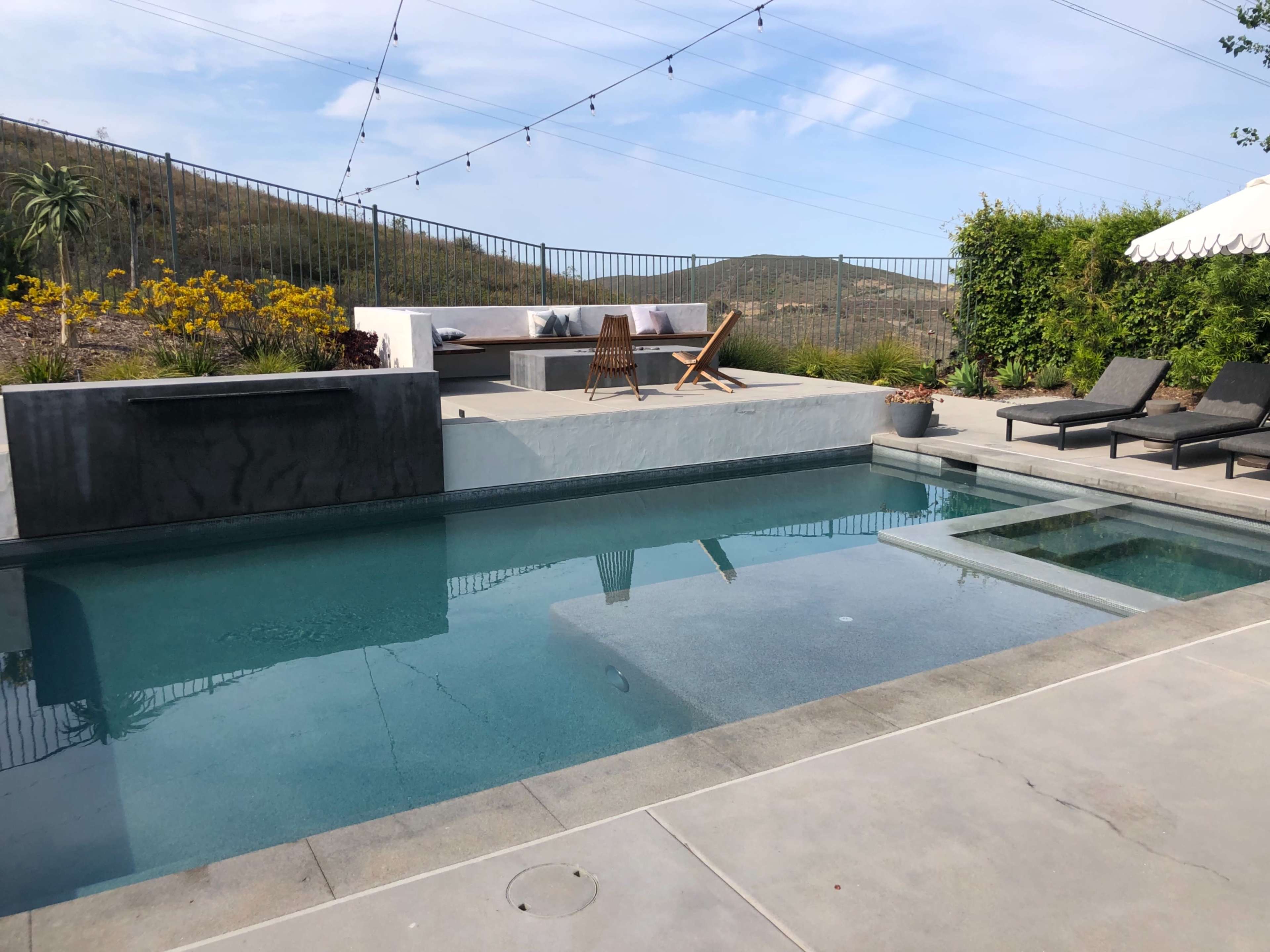 The image shows a modern backyard with a swimming pool, lounge chairs, and a seating area against a backdrop of hills and a blue sky.