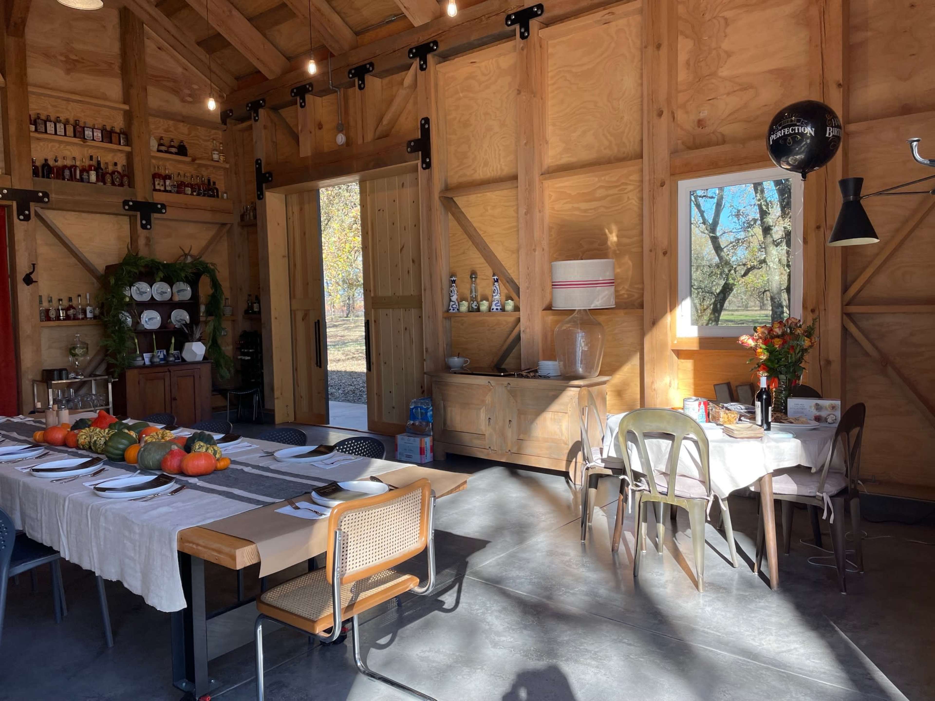 The image shows the interior of a rustic wooden dining space featuring two tables set with dishes and decorative items, along with a bar area stocked with bottles.