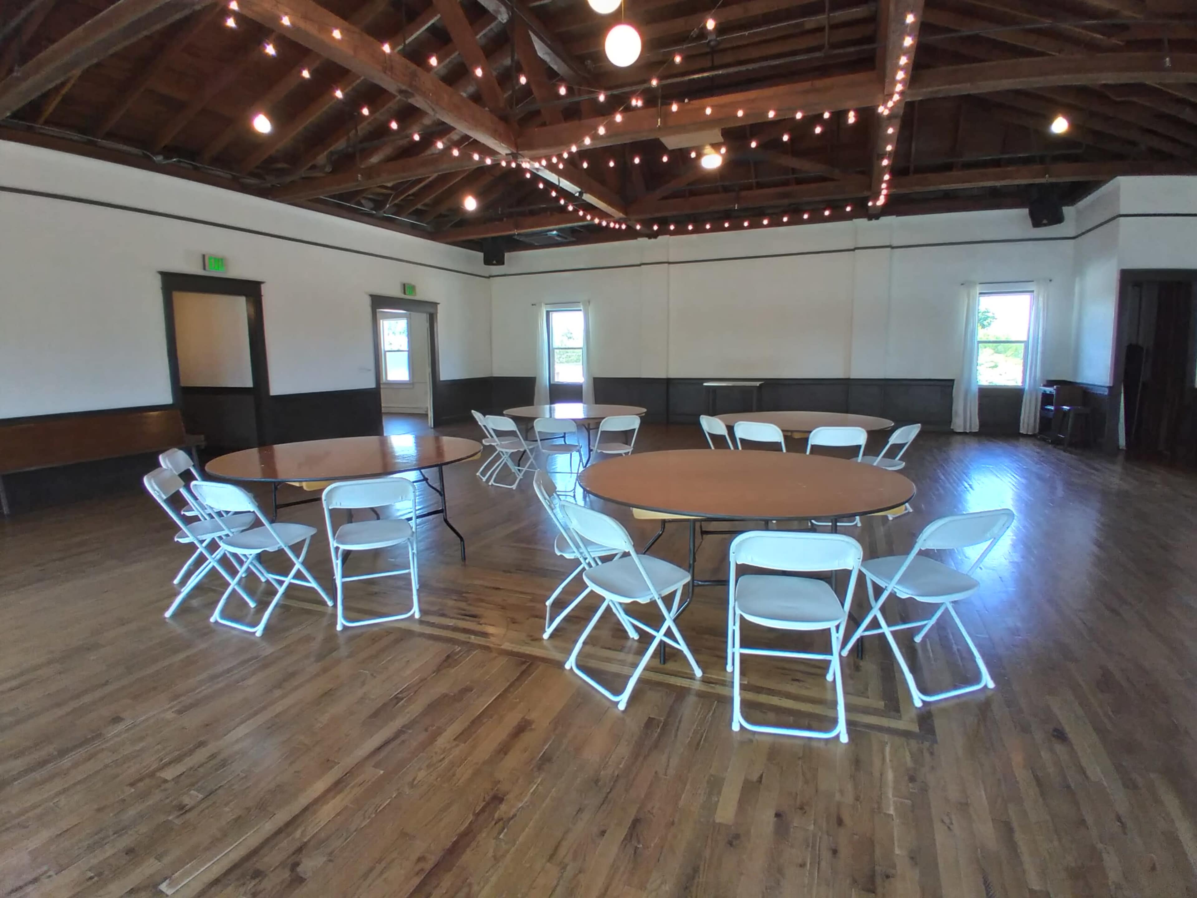 A large, well-lit room with wooden flooring, featuring several round tables surrounded by white chairs and string lights overhead.