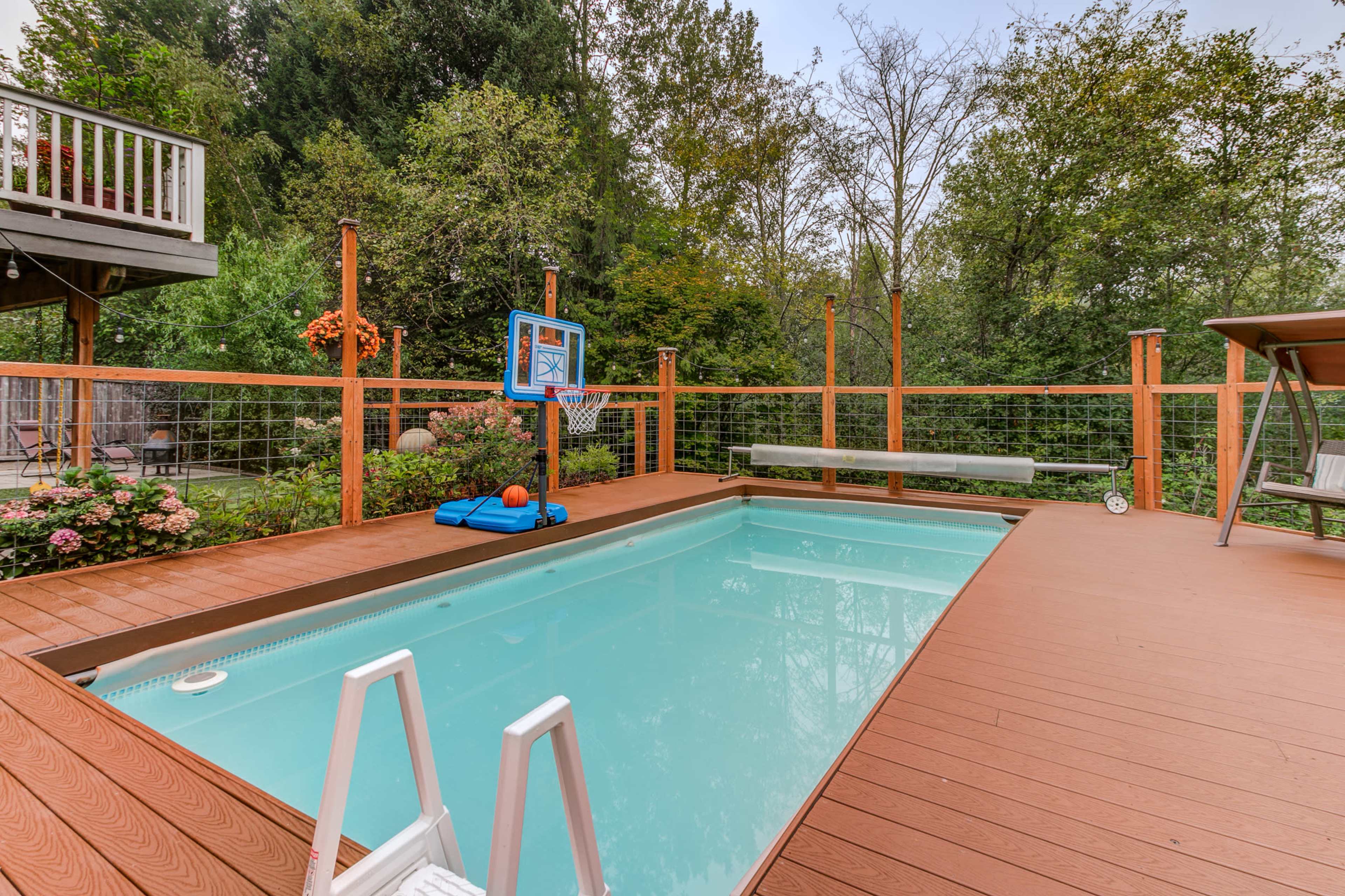 The image shows a backyard with a swimming pool surrounded by a wooden deck, a basketball hoop, and lush greenery in the background.