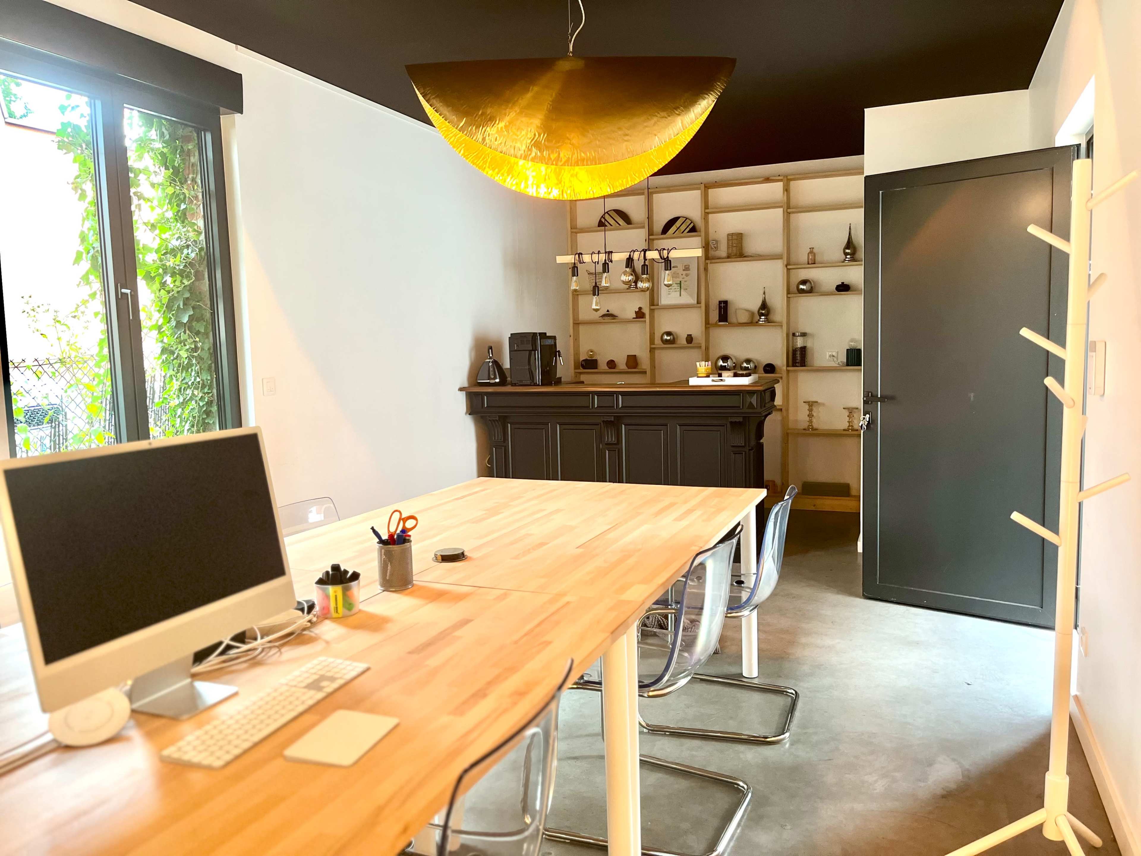 The image shows a modern workspace with a wooden table and clear chairs, featuring a computer, and a dark-colored shelf in the background displaying various items.
