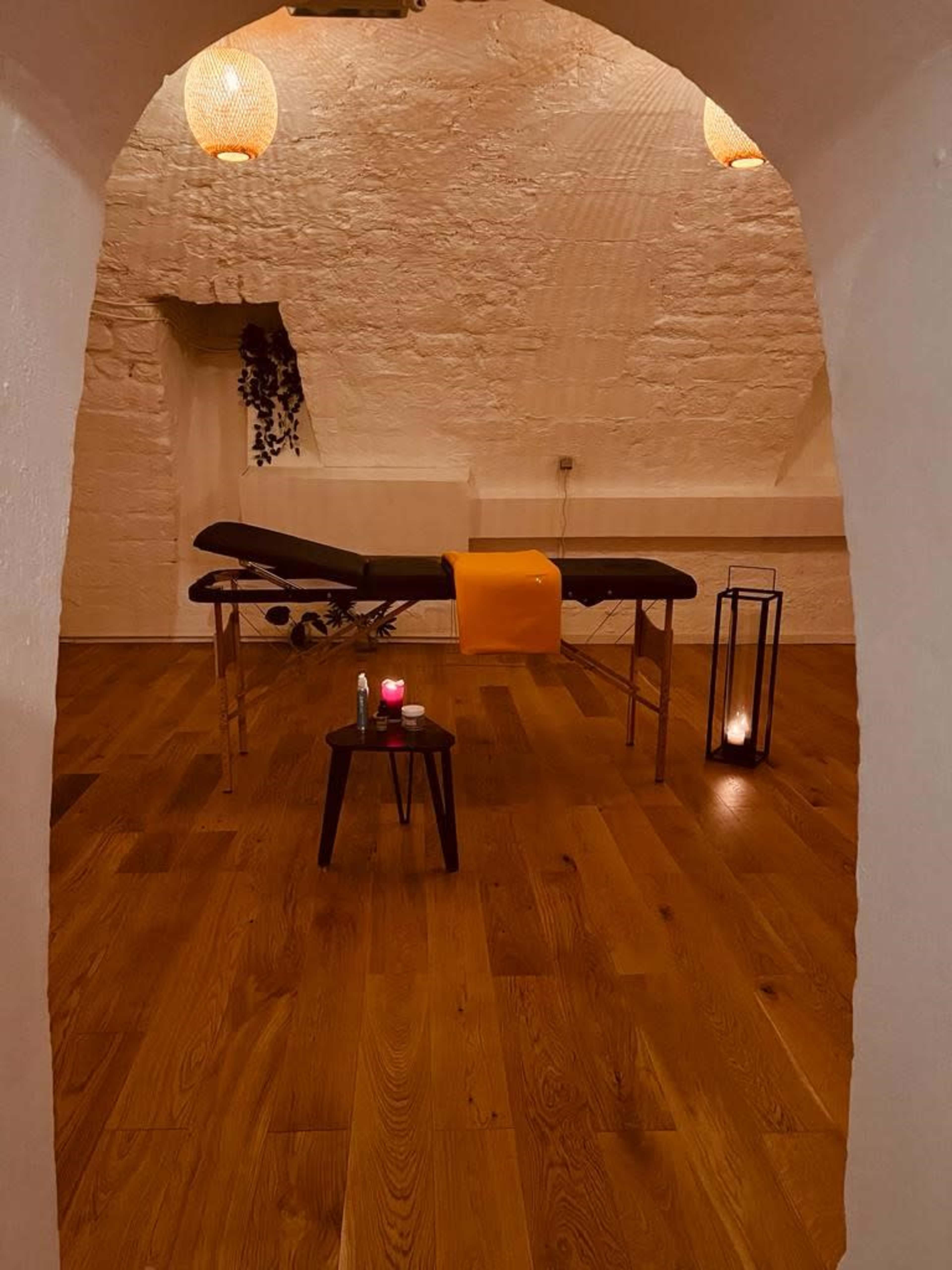 A massage room features a treatment table with an orange towel, surrounded by candles and wooden flooring.
