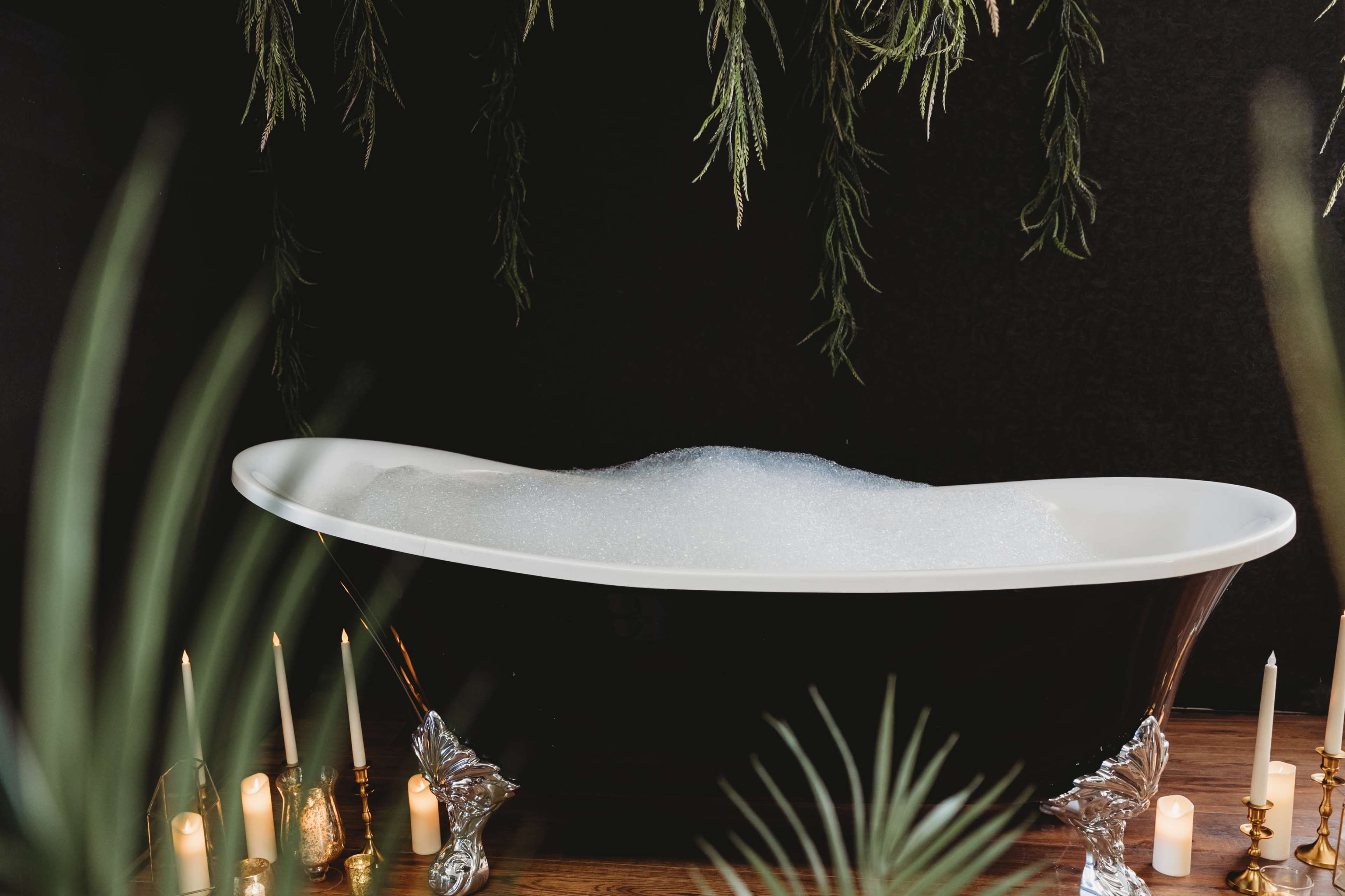 A freestanding white bathtub filled with bubbles is set against a dark background, surrounded by greenery and candles.
