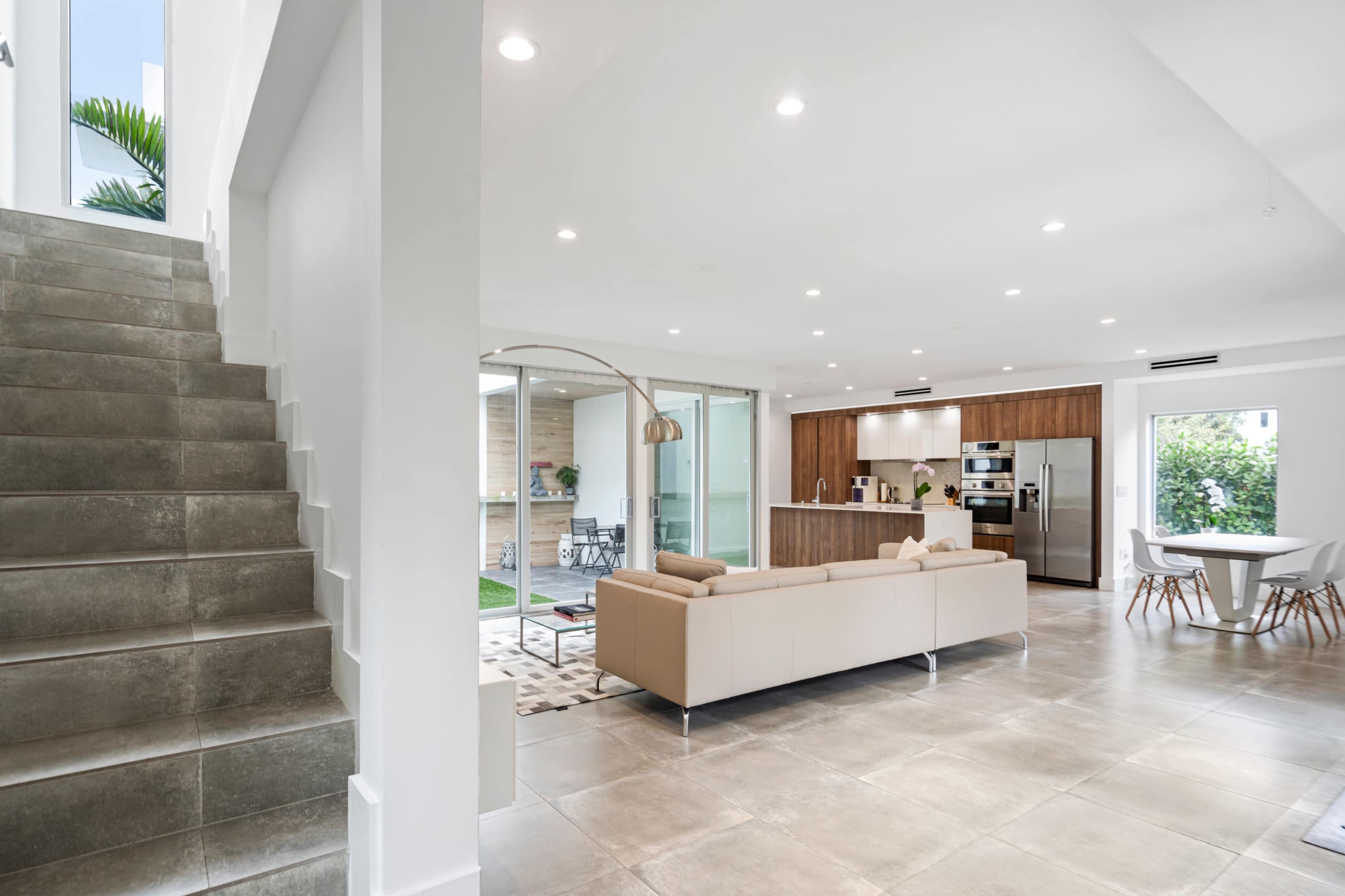 A modern open-concept living space features a staircase, a beige sofa, and a kitchen with wooden cabinets and stainless steel appliances.