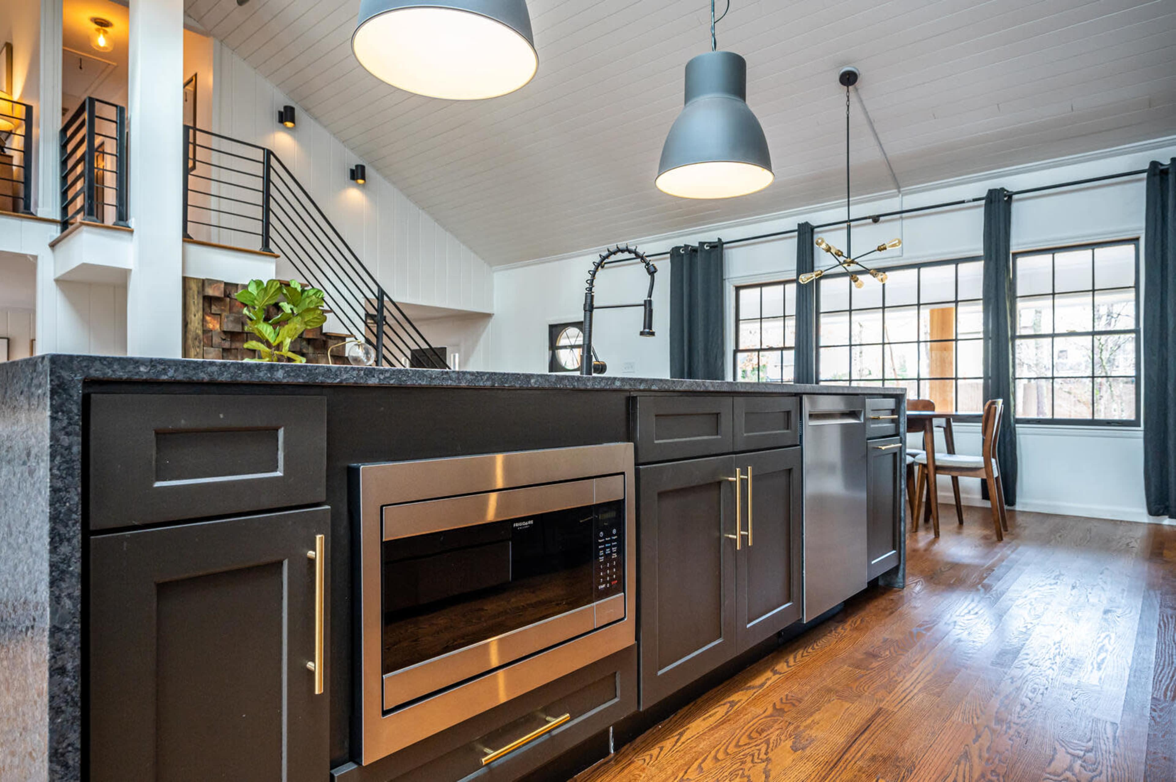 A modern kitchen features dark cabinets, a built-in microwave, and pendant lighting, with an open layout leading to a dining area and staircase.