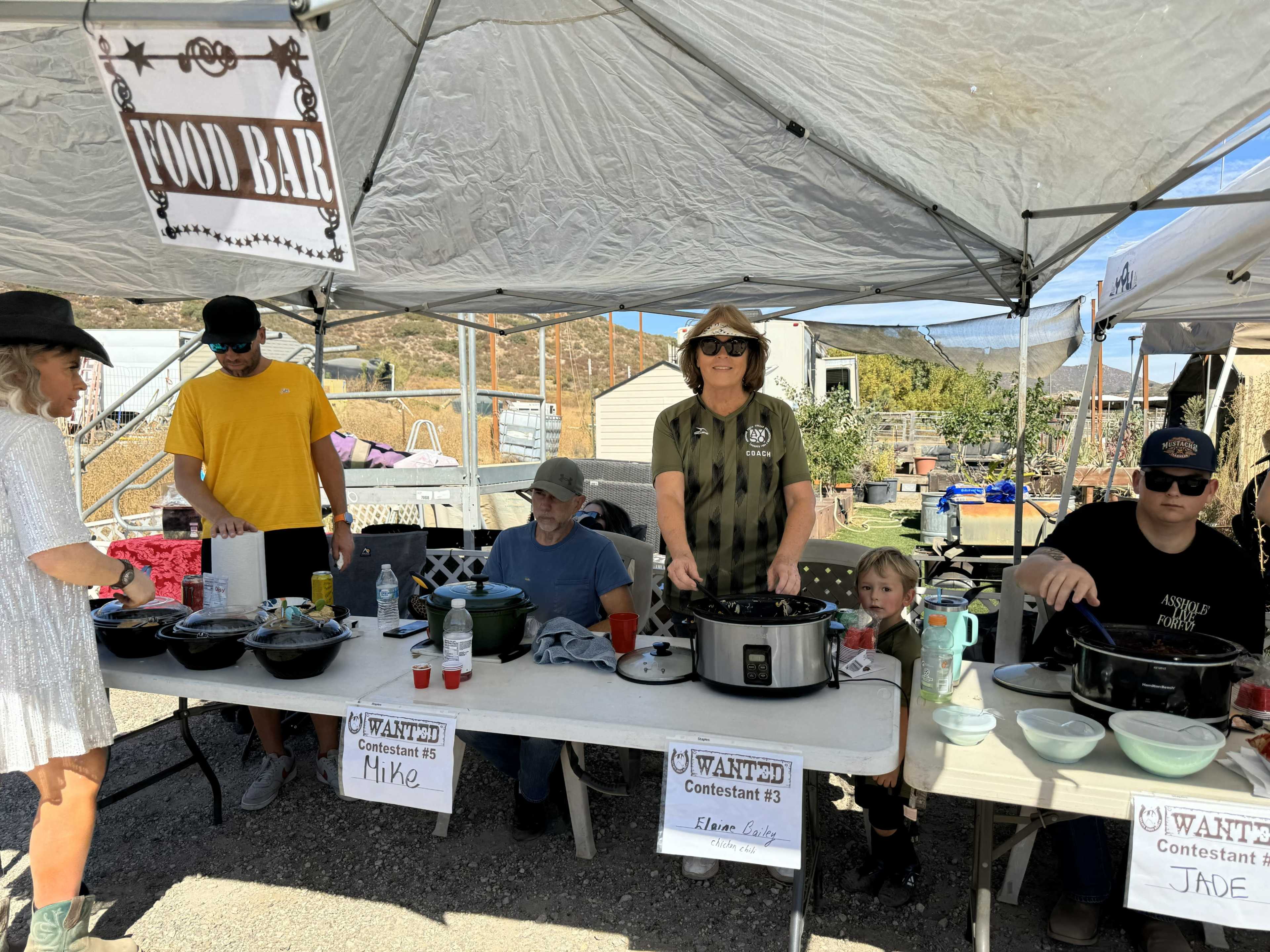 A group of people is gathered around a food setup at an outdoor event, with several tables displaying dishes and signs indicating contestants.