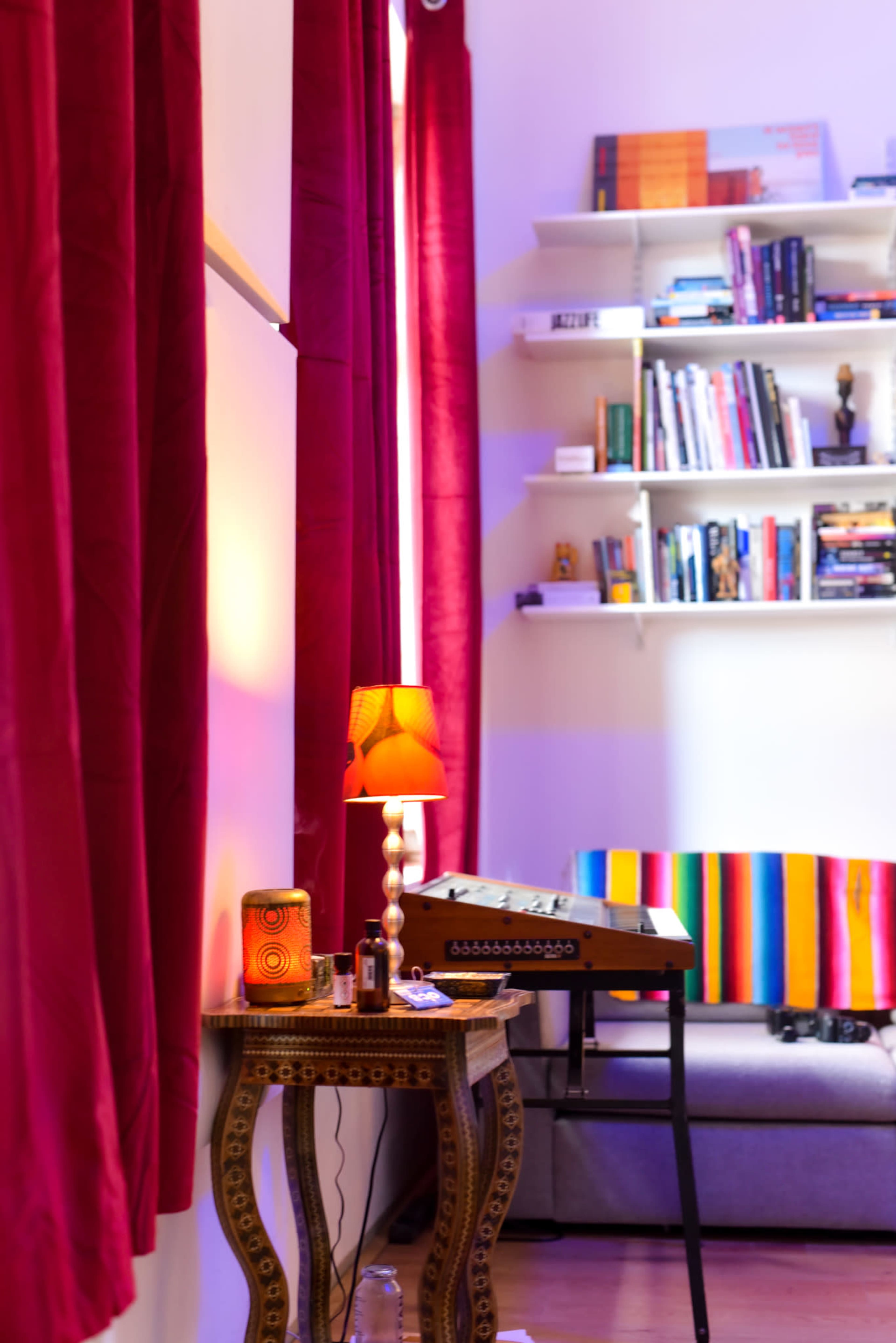 A cozy room features a decorated sofa with a colorful throw and a small table with a lamp, surrounded by red curtains and a shelf filled with books.