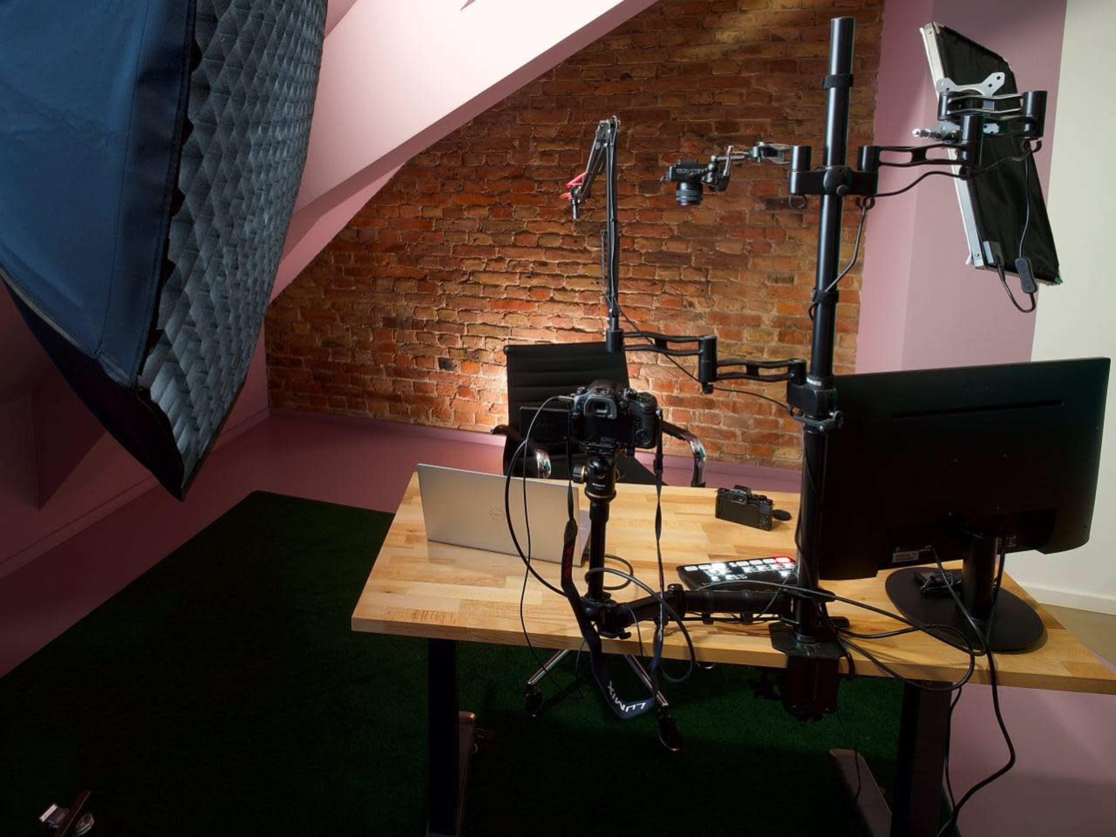 A workspace set up with a wooden desk, a laptop, dual monitors, a camera mounted on a stand, and professional lighting against a brick wall.