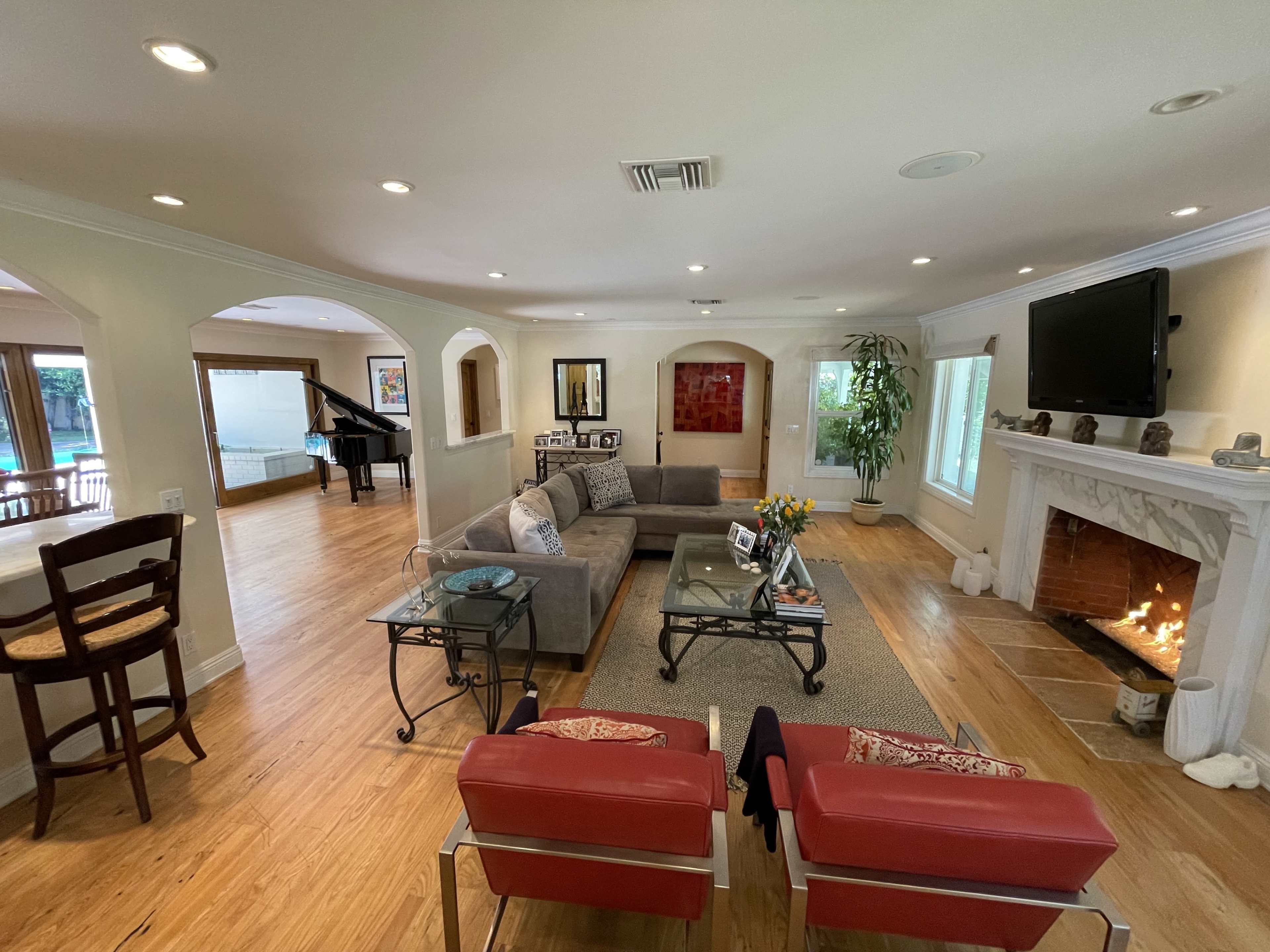 A spacious living room with a gray sectional sofa, a glass coffee table, and a fireplace, complemented by an adjacent area featuring a piano and large windows.