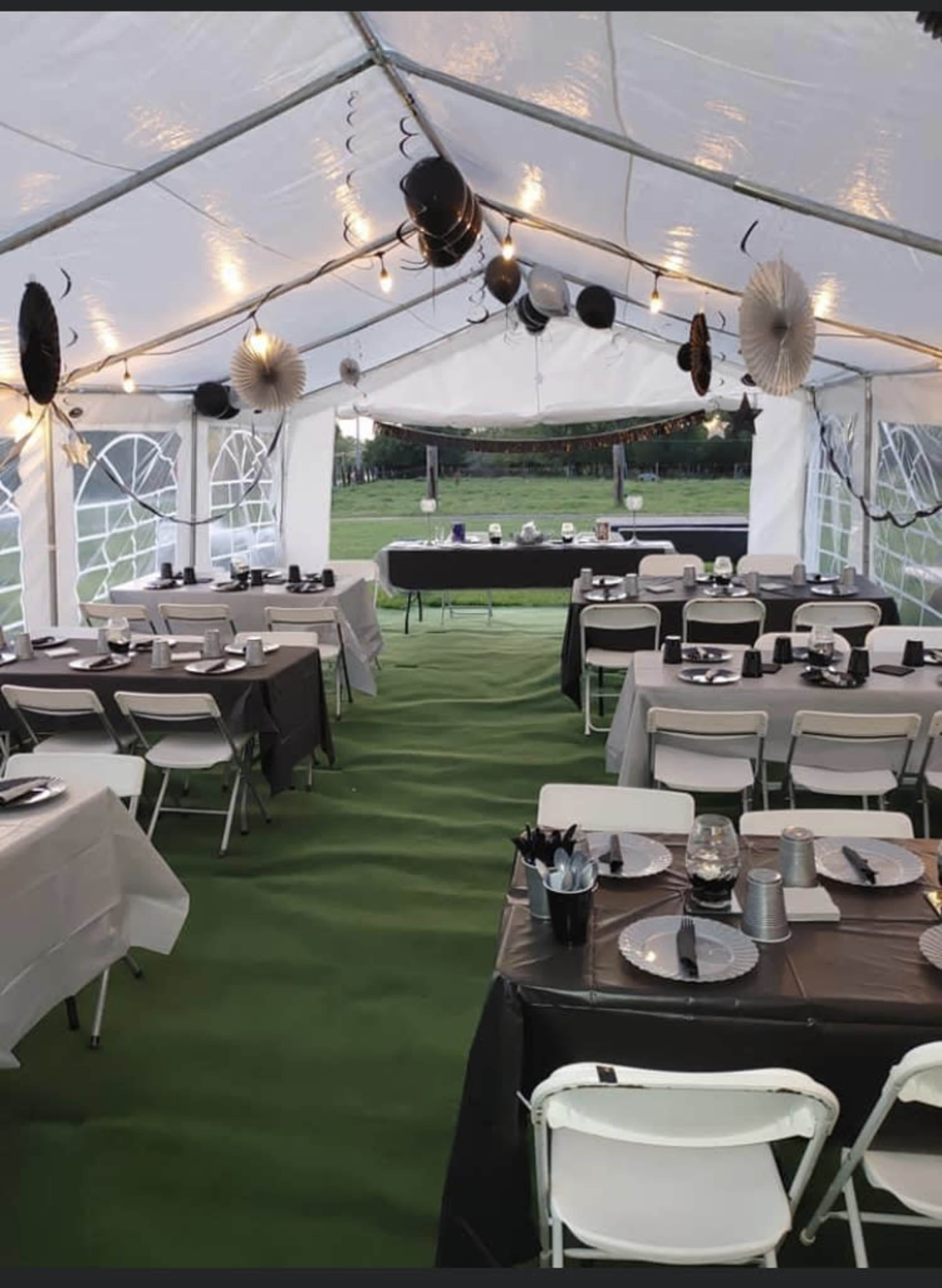 A tent is set up with tables covered in black and white linens, arranged with plates and utensils, and decorated with hanging paper decorations and string lights.