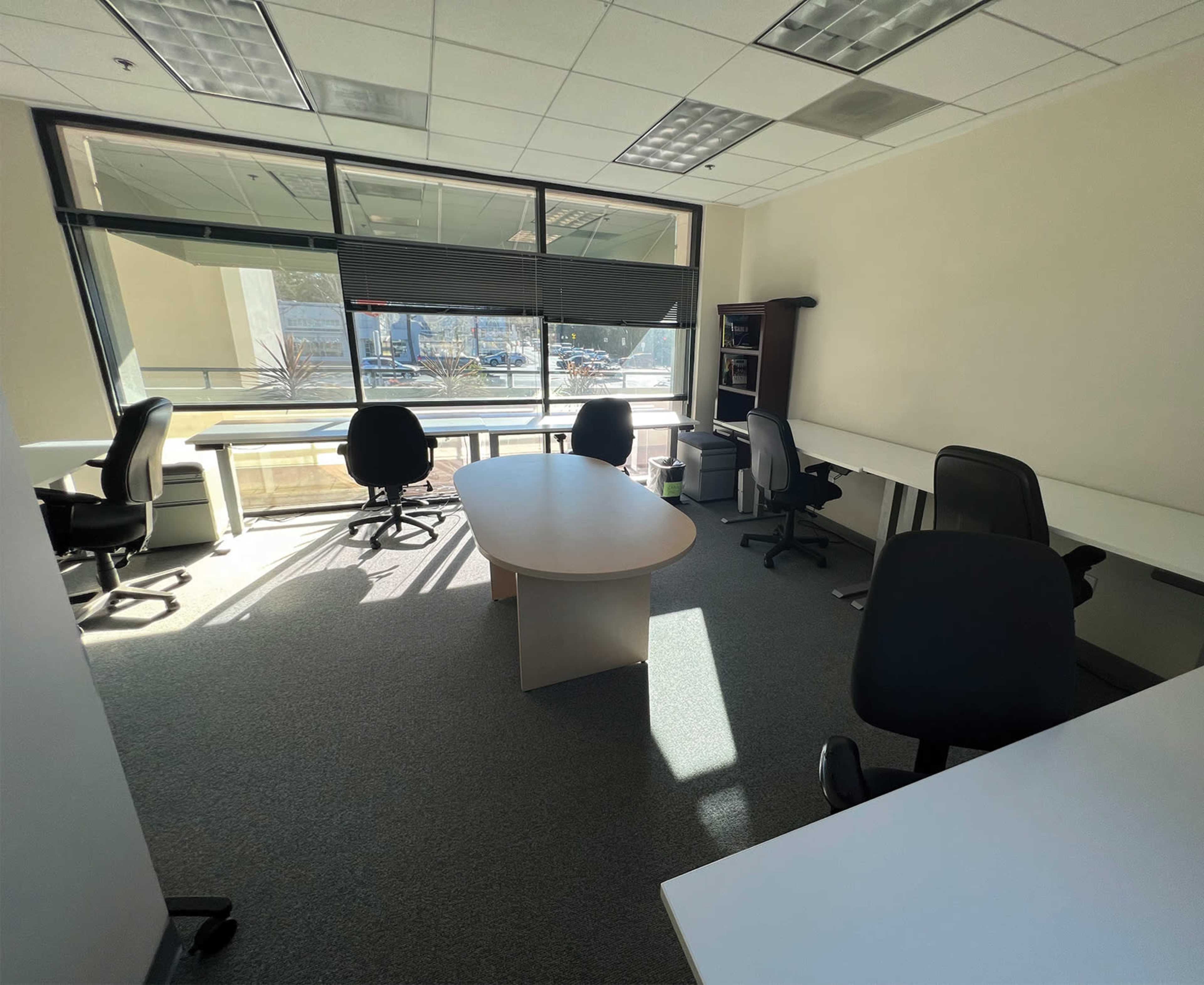A bright office space featuring several desks and chairs, with large windows providing a view outside.