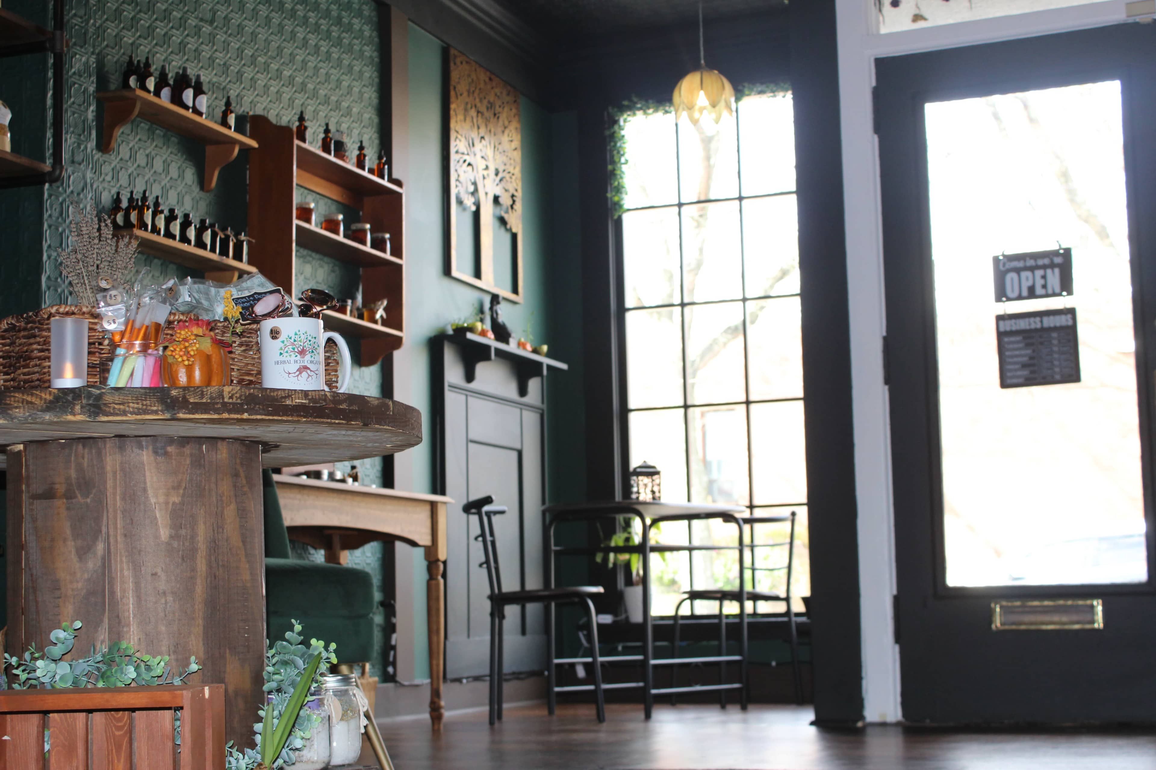 A cozy interior of a café featuring wooden furniture, green walls, shelves with jars, and a large window with an open sign.