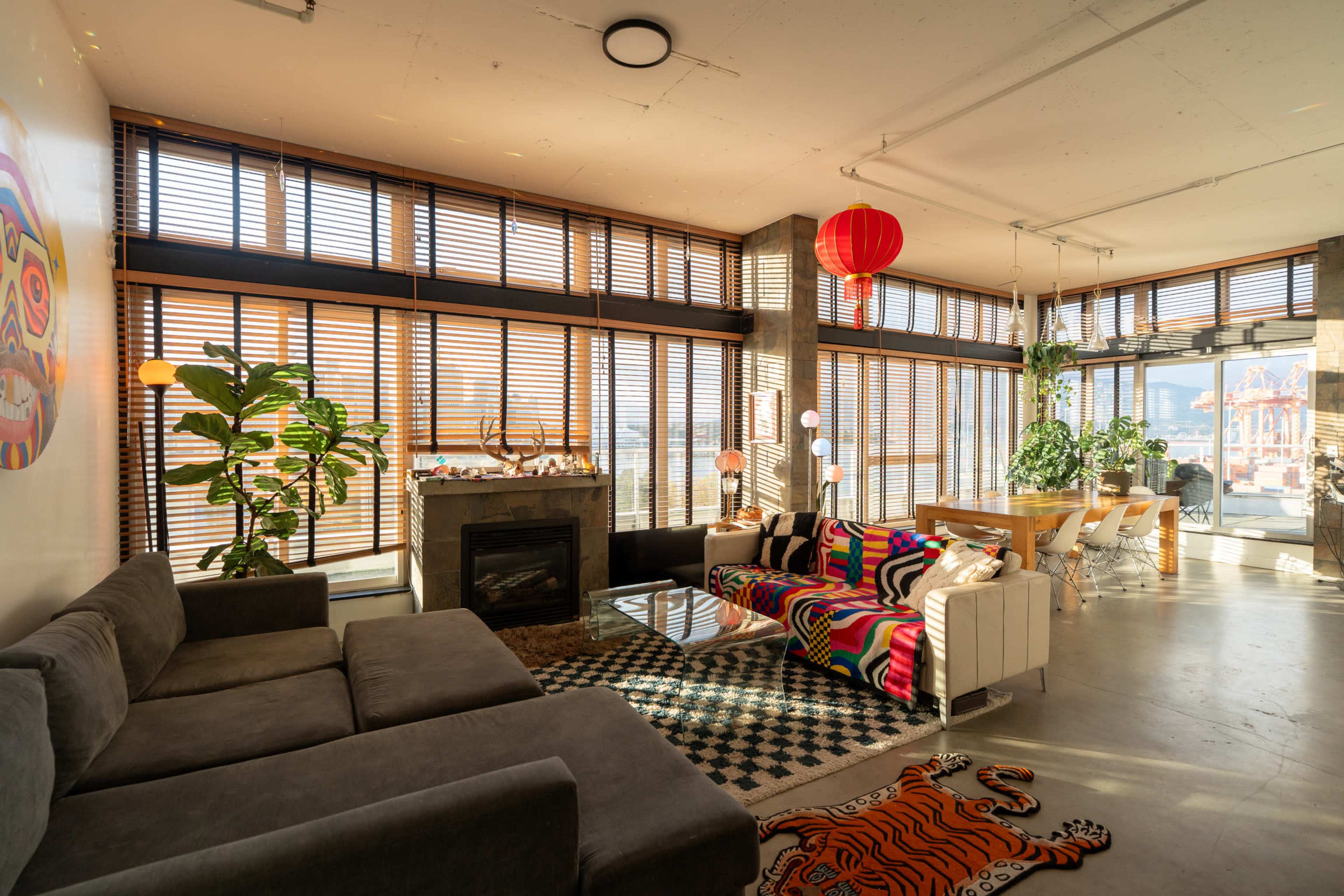 The image shows a modern living space featuring a gray sectional sofa, colorful patterned furniture, large windows with wooden blinds, and an open dining area with a view.
