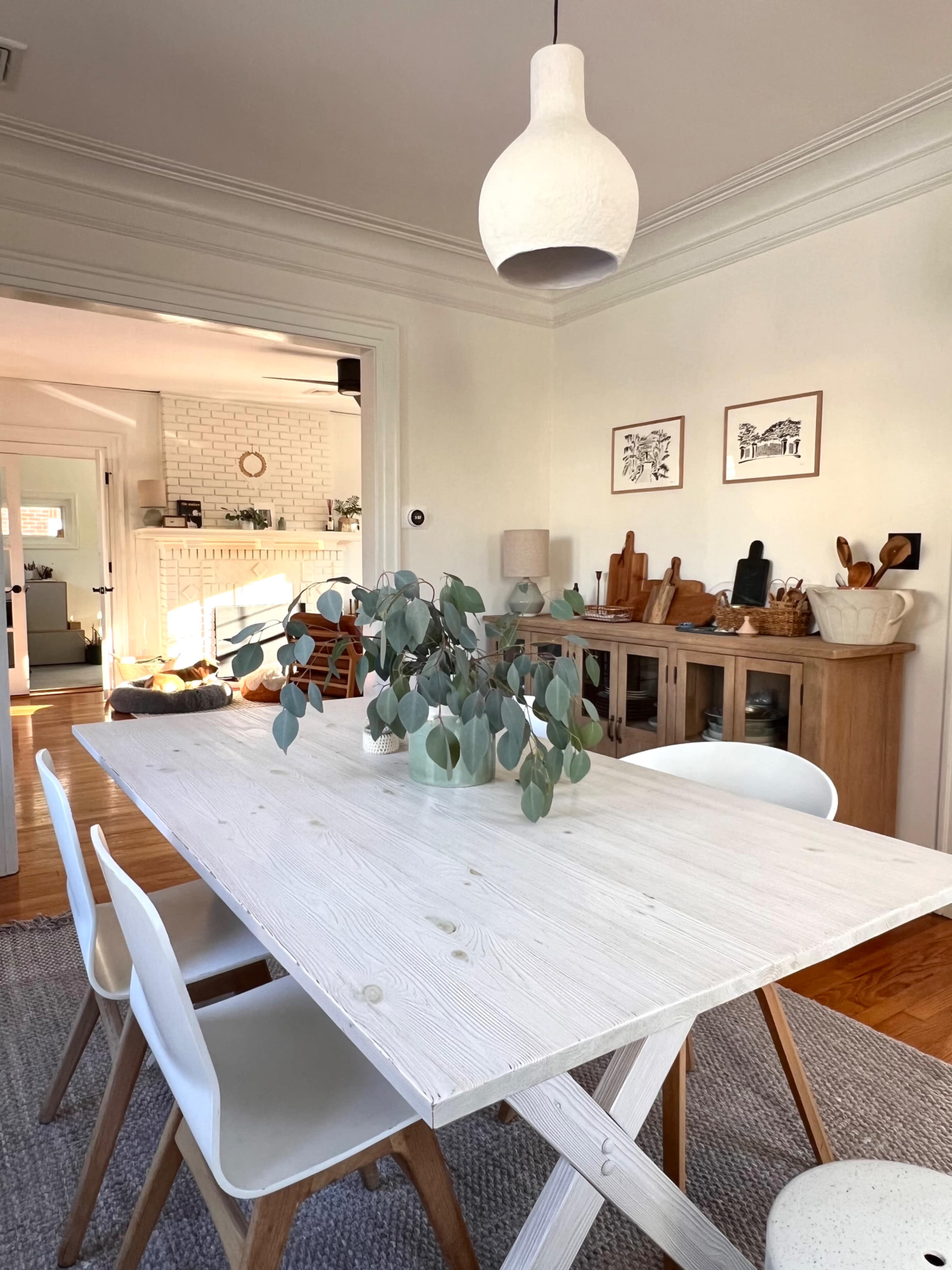 A dining table with a white surface and several chairs is positioned in a room with a light fixture and decorative elements in the background.