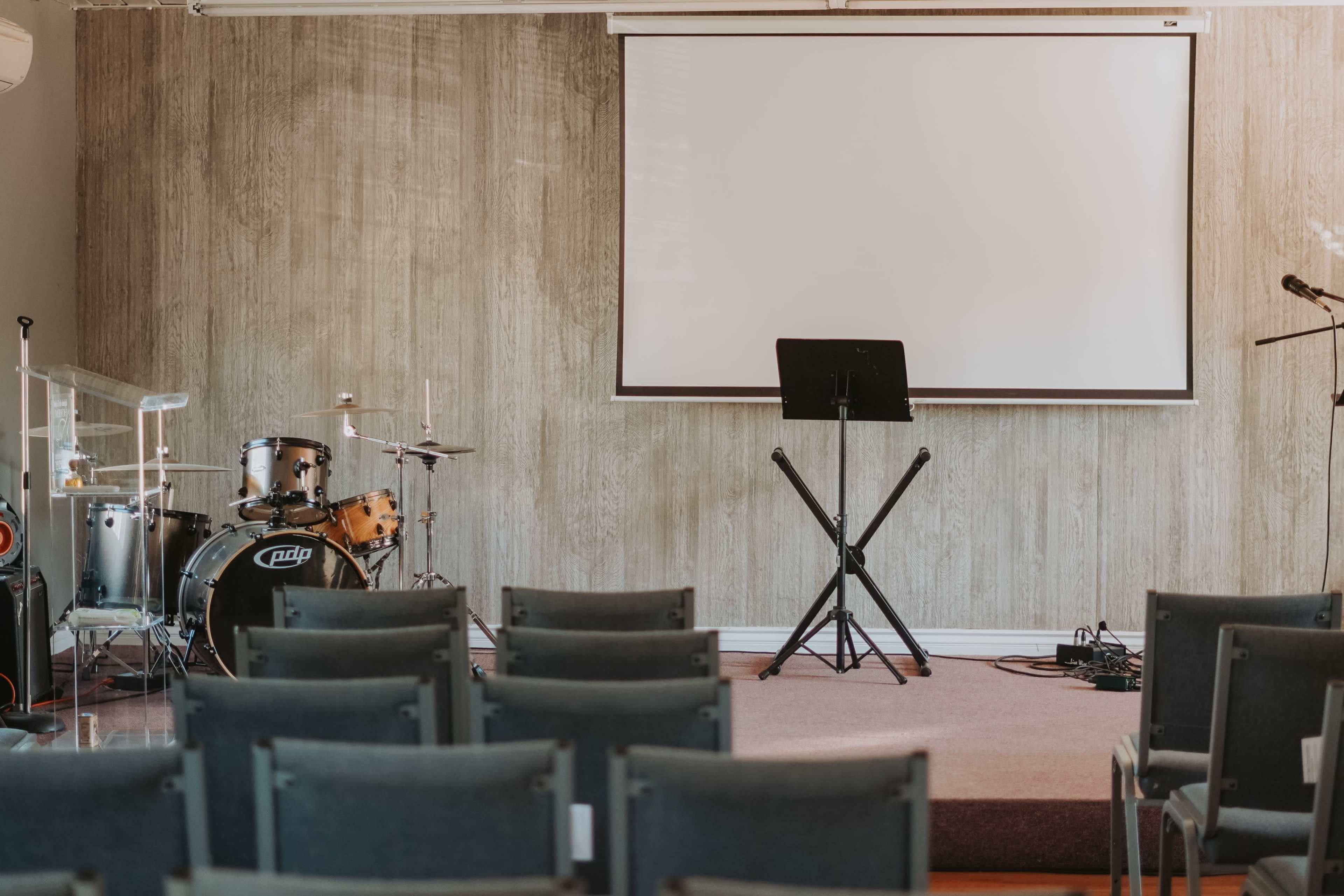A stage setup in a room with a blank screen, musical instruments, and chairs arranged in rows.