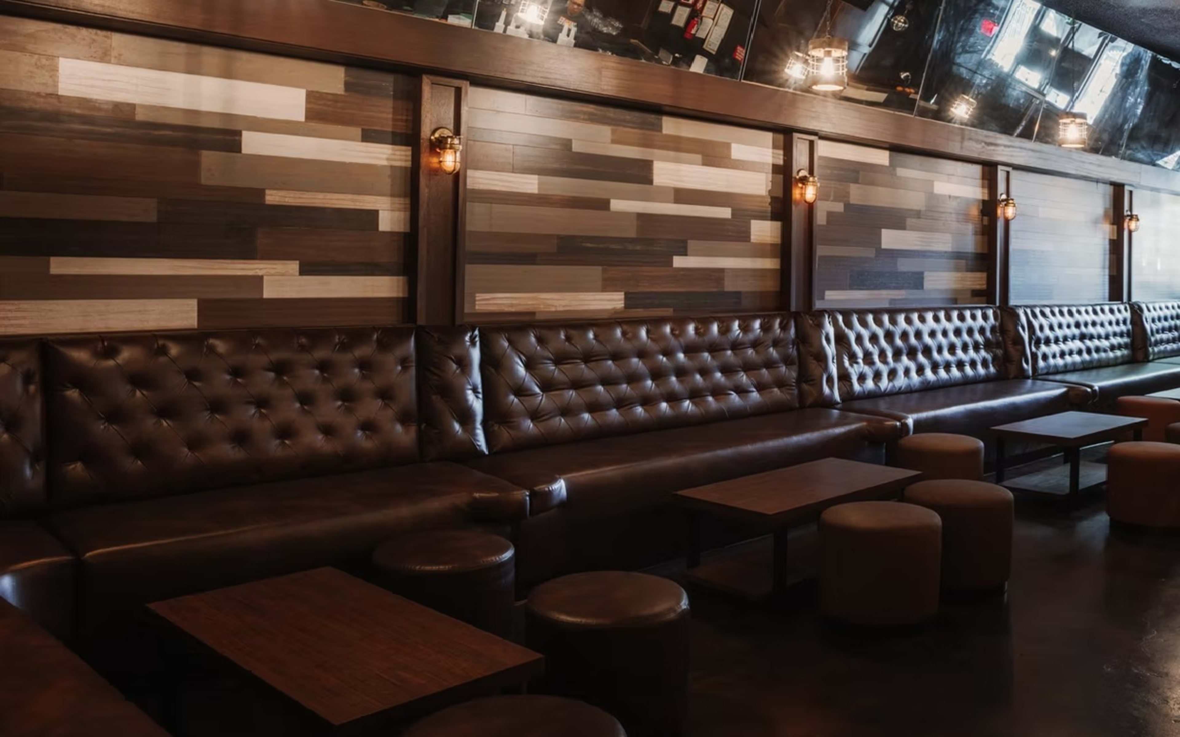 The image shows a dimly lit lounge area with a leather tufted seating arrangement against a wooden wall featuring a variety of wood panel designs.