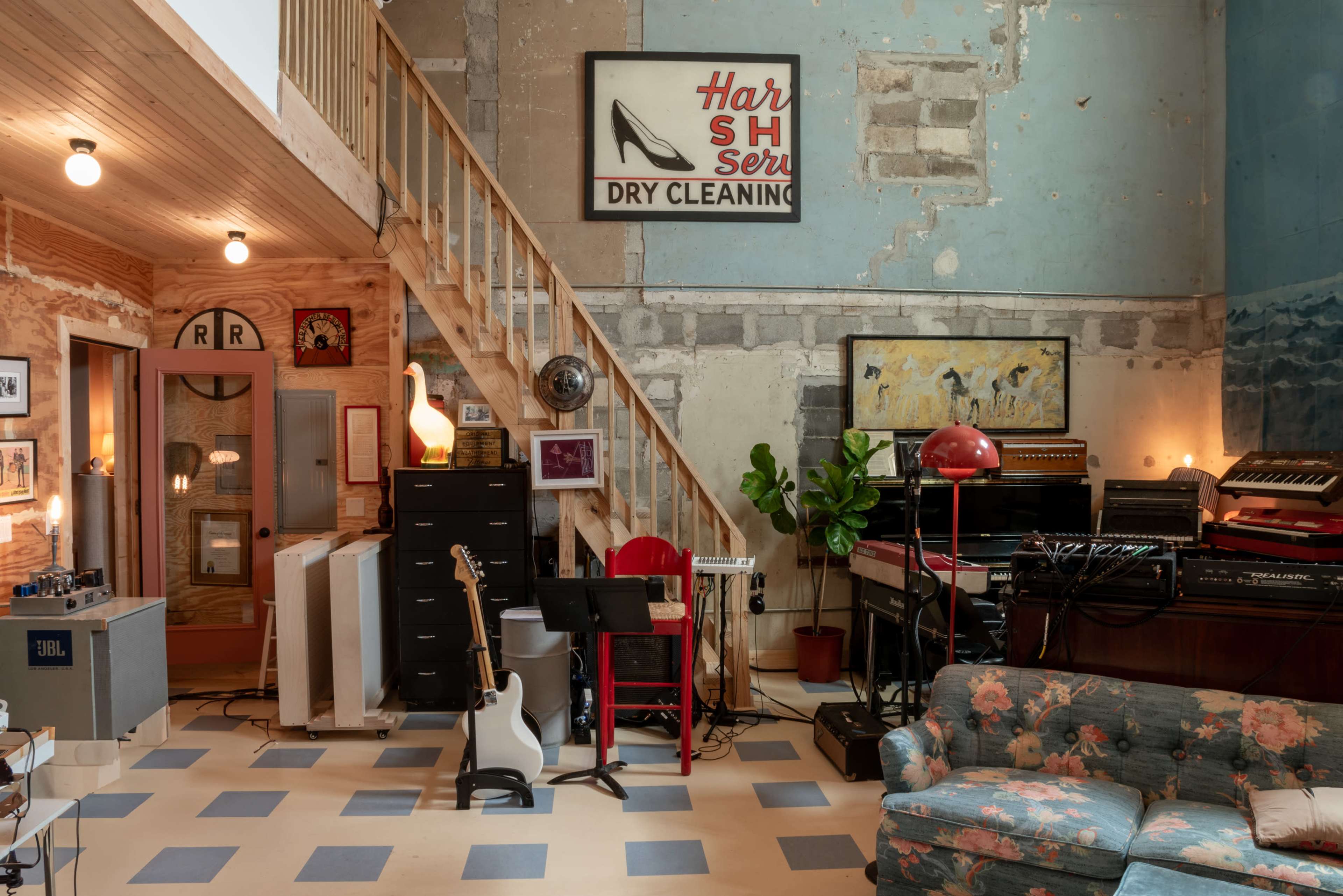 The image depicts an eclectic interior of a studio or workshop featuring a staircase, vintage furniture, musical equipment, and artwork on the walls.