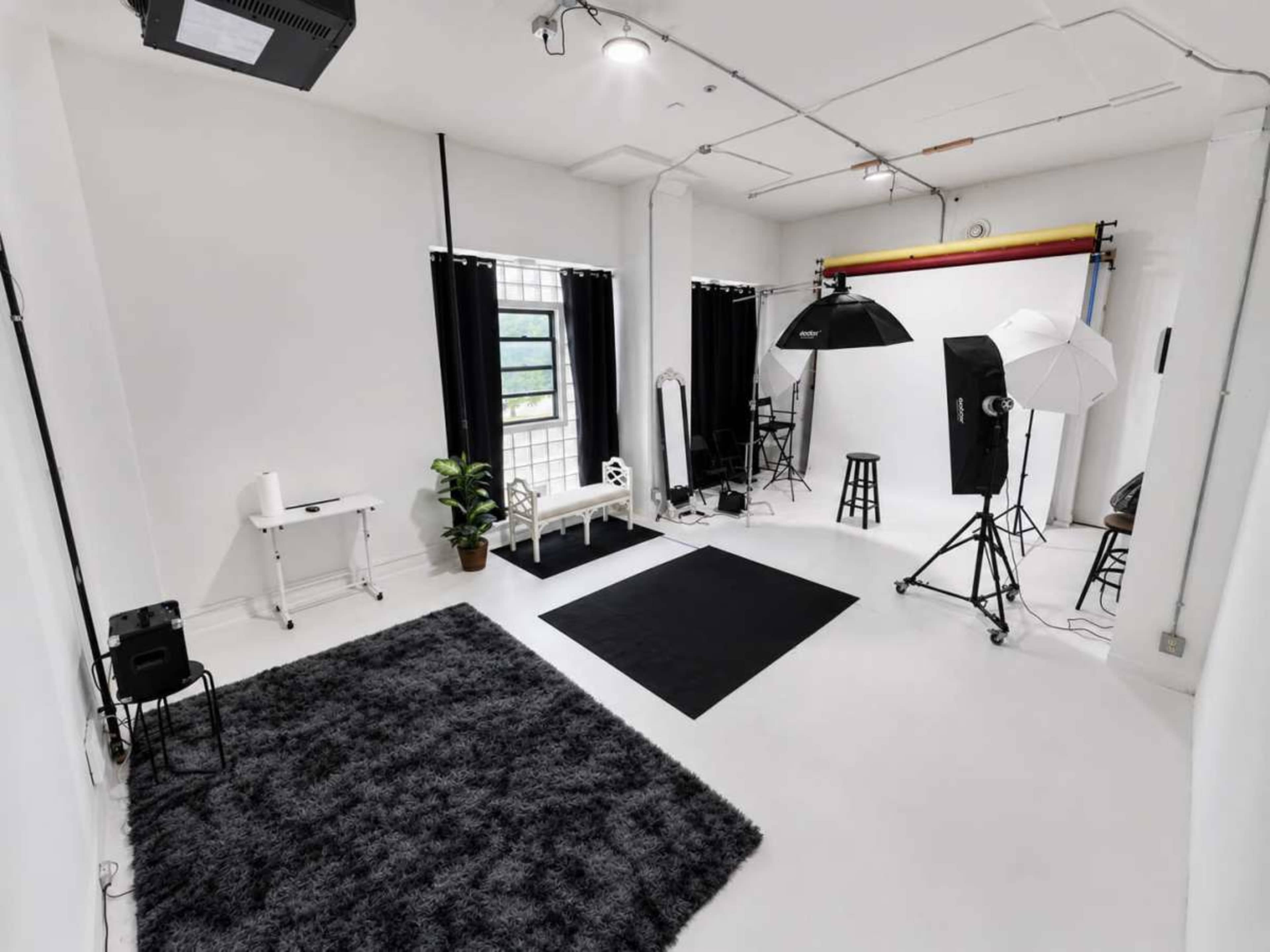 The image shows a well-lit photography studio featuring various equipment, including softbox lights, a black rug, and a small white seating area.