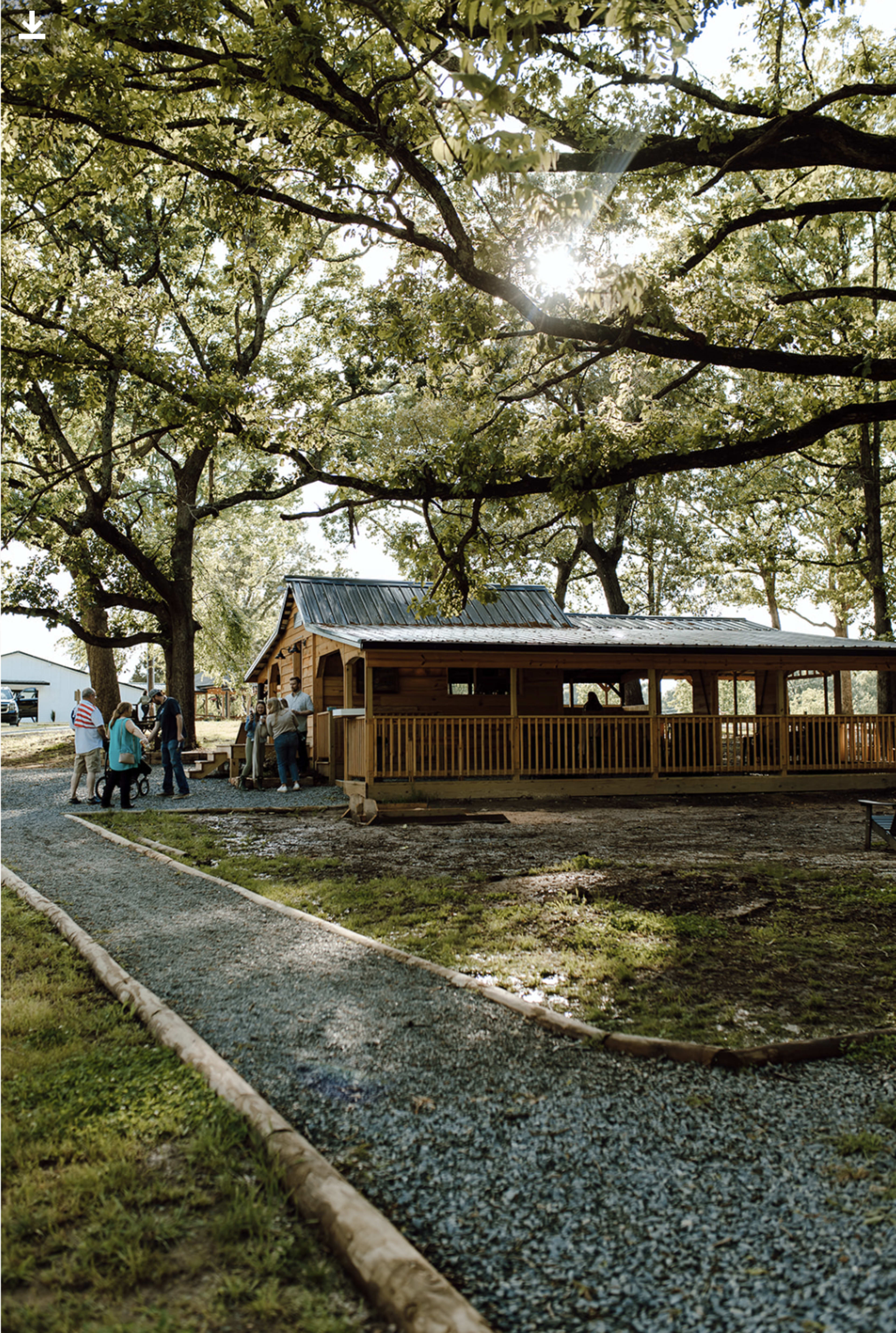 A wooden building with a porch stands under large trees, while a group of people gathers nearby on a gravel pathway.