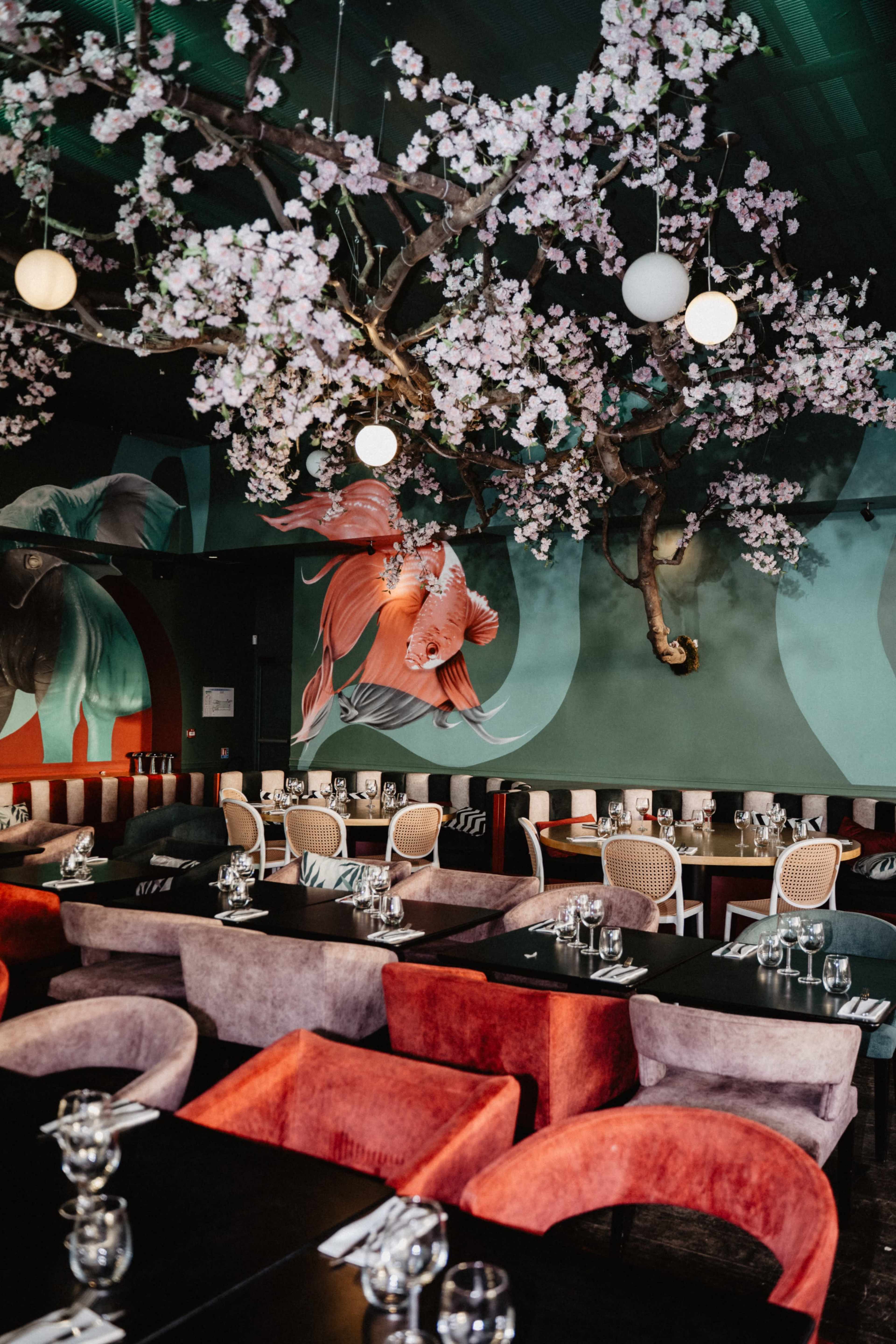 The image depicts a modern restaurant interior featuring elegantly arranged tables with colorful chairs, adorned by a large cherry blossom tree and vibrant wall murals of goldfish.
