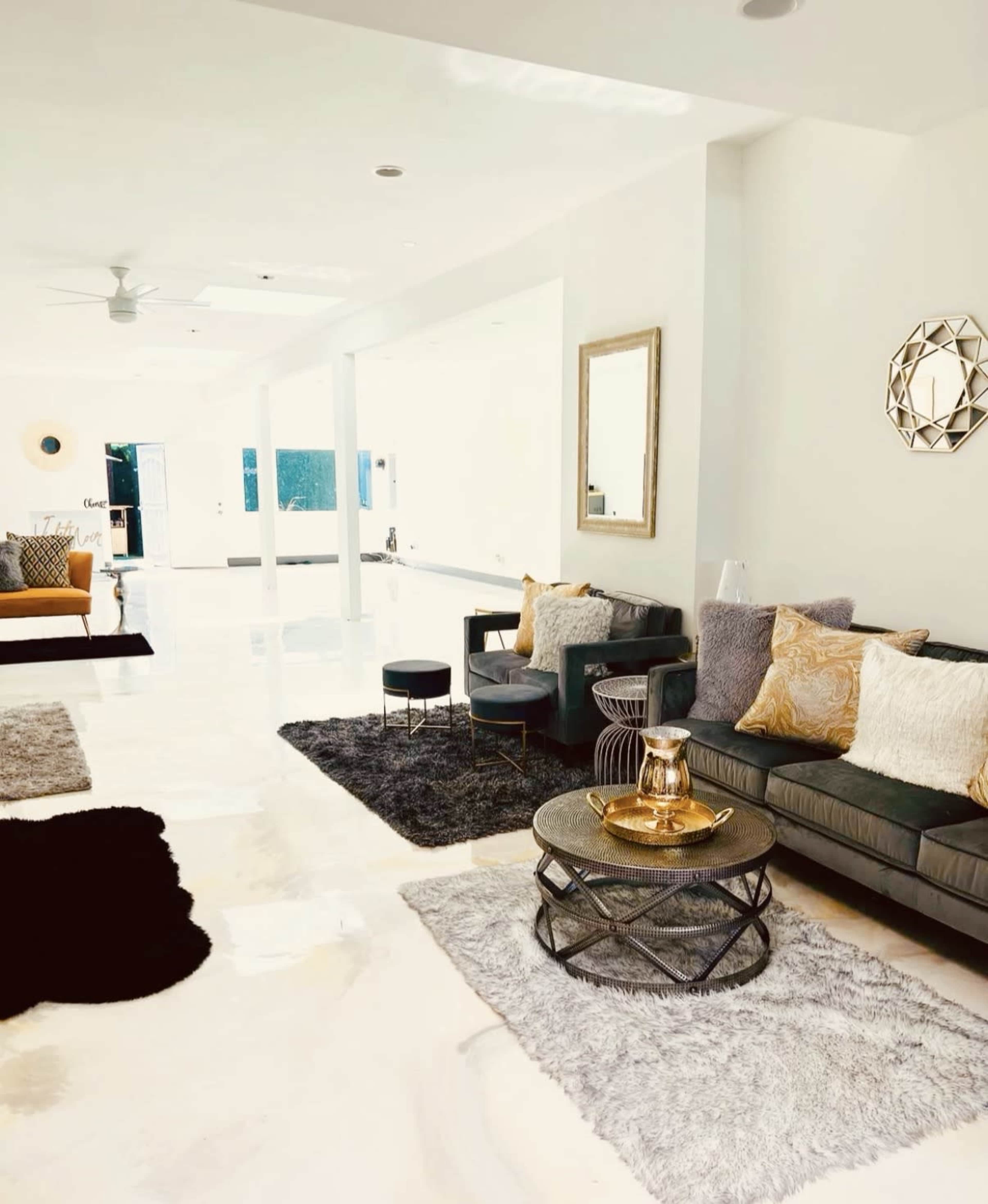 The image shows a bright and modern living room with a light-colored floor, a dark sectional sofa, decorative pillows, a coffee table with a teapot, and a large mirror on the wall.