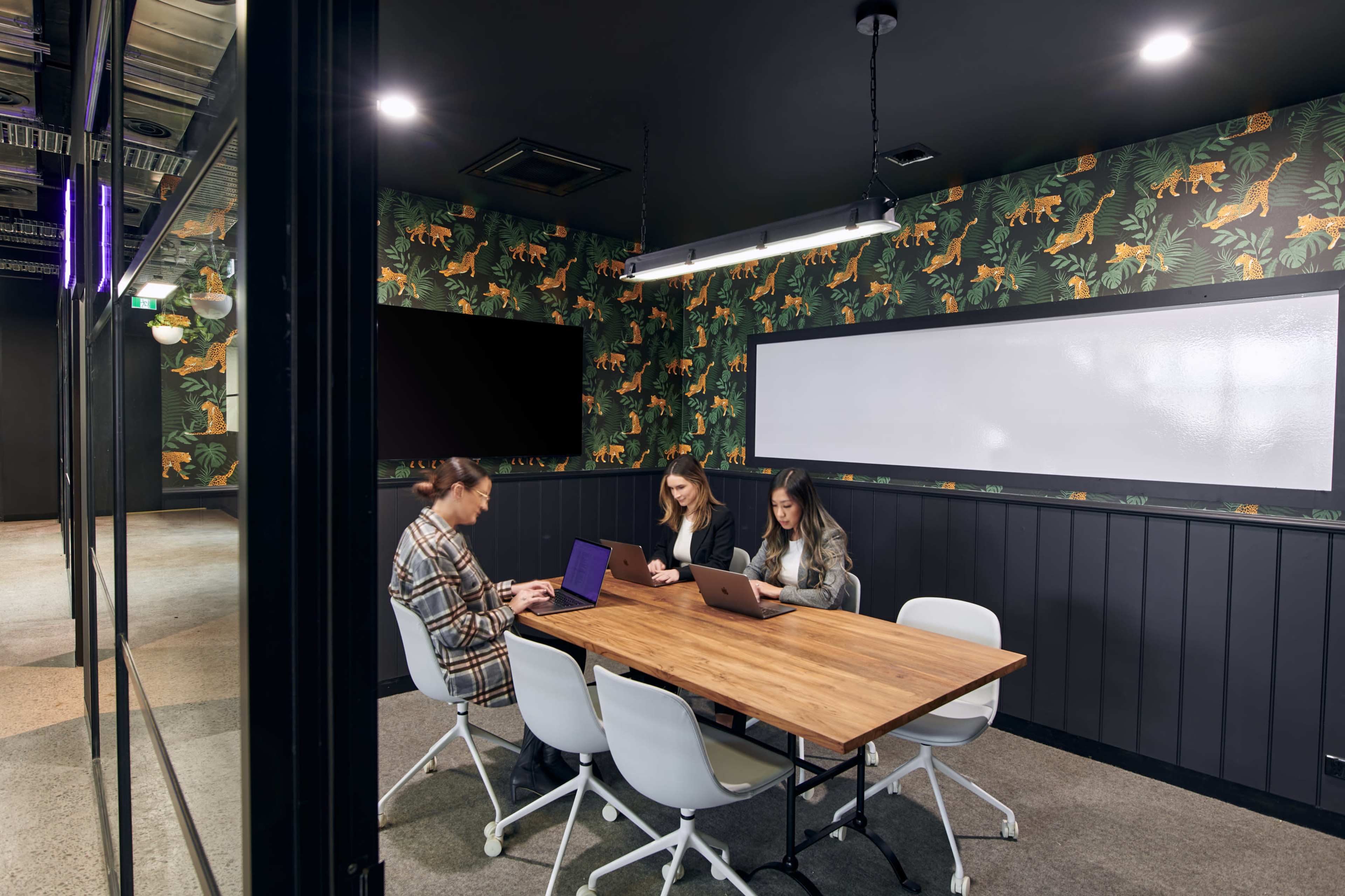 Three people sit at a wooden table in a modern conference room with a jungle-themed wallpaper.