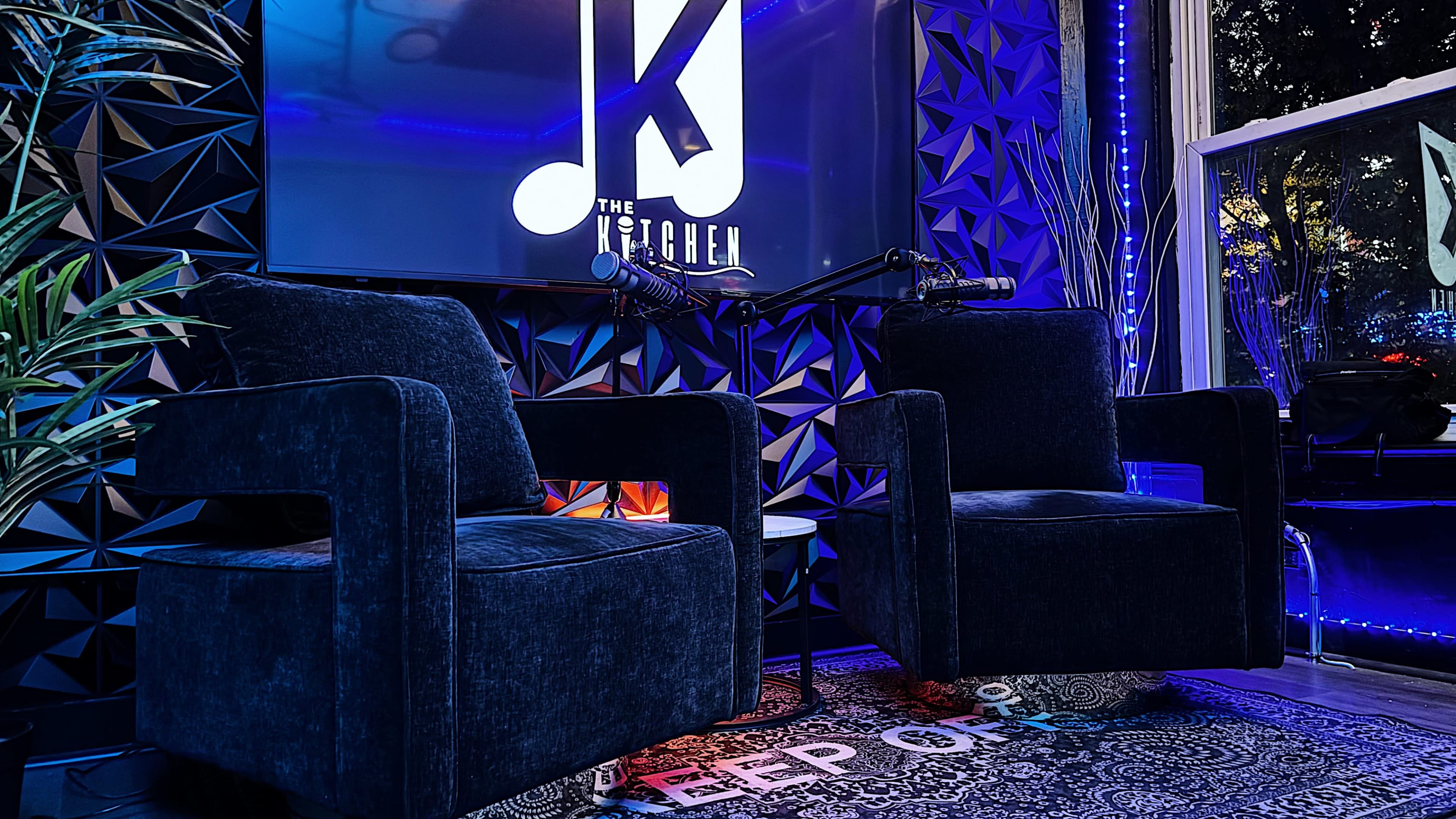 The image shows a stylish studio setup featuring two blue chairs, a decorative rug, and a large screen displaying a logo, all with ambient lighting.