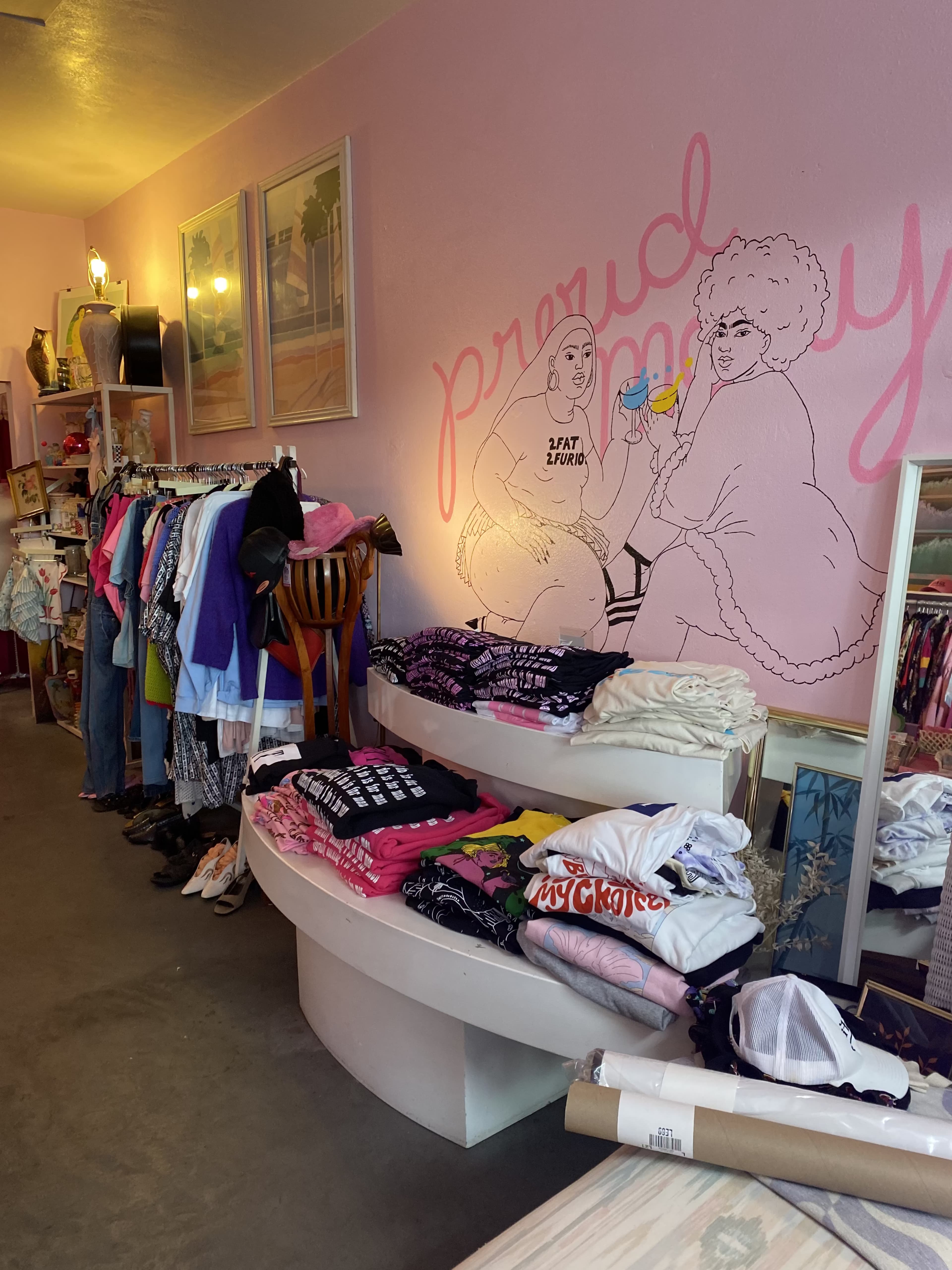 A clothing store interior featuring racks of apparel on one side and curved tables displaying folded shirts on the other, with a pink mural on the wall depicting two figures.