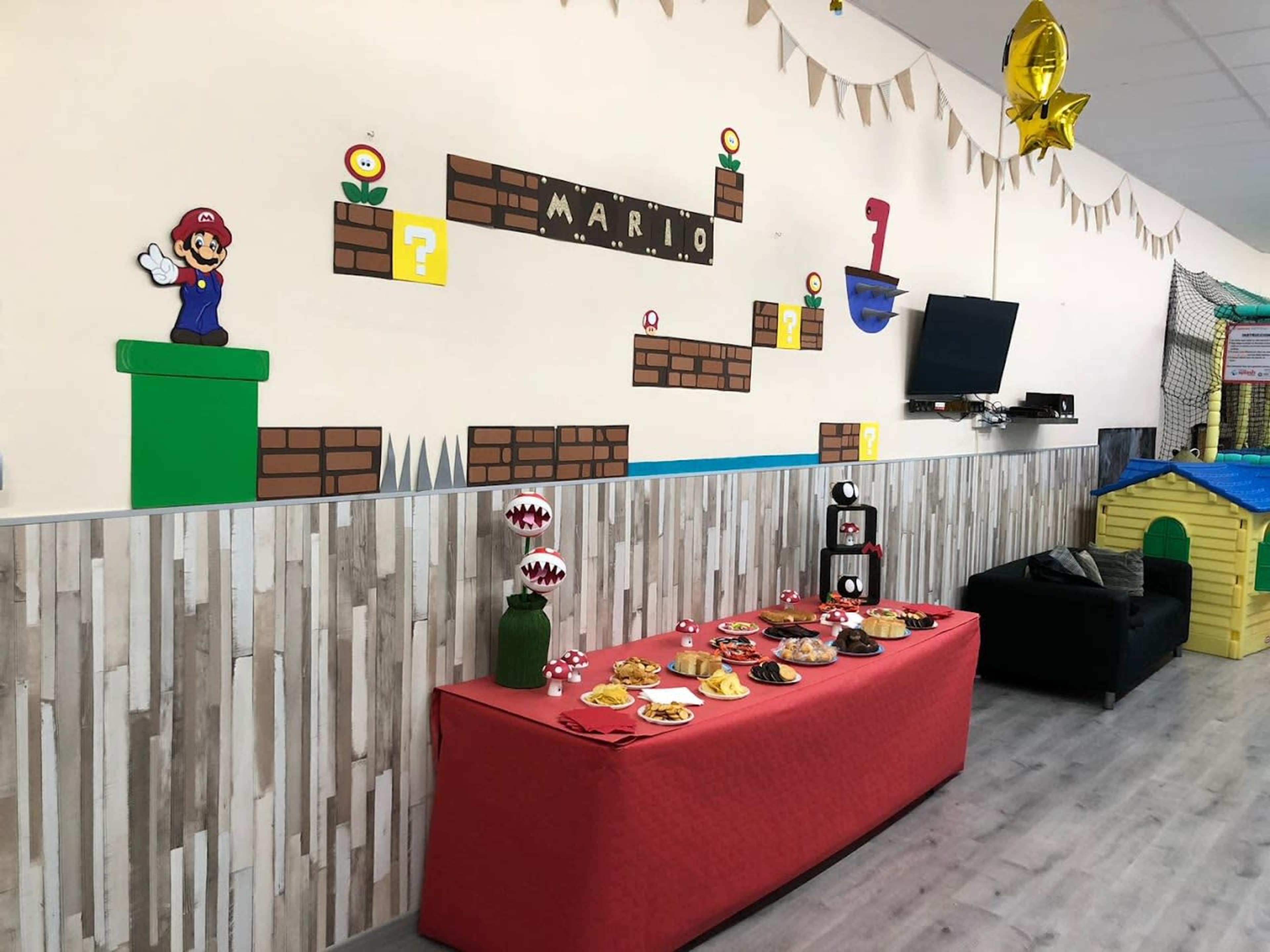 A decorated room features a Mario-themed wall display with game characters and blocks, alongside a table set with snacks and a black couch.