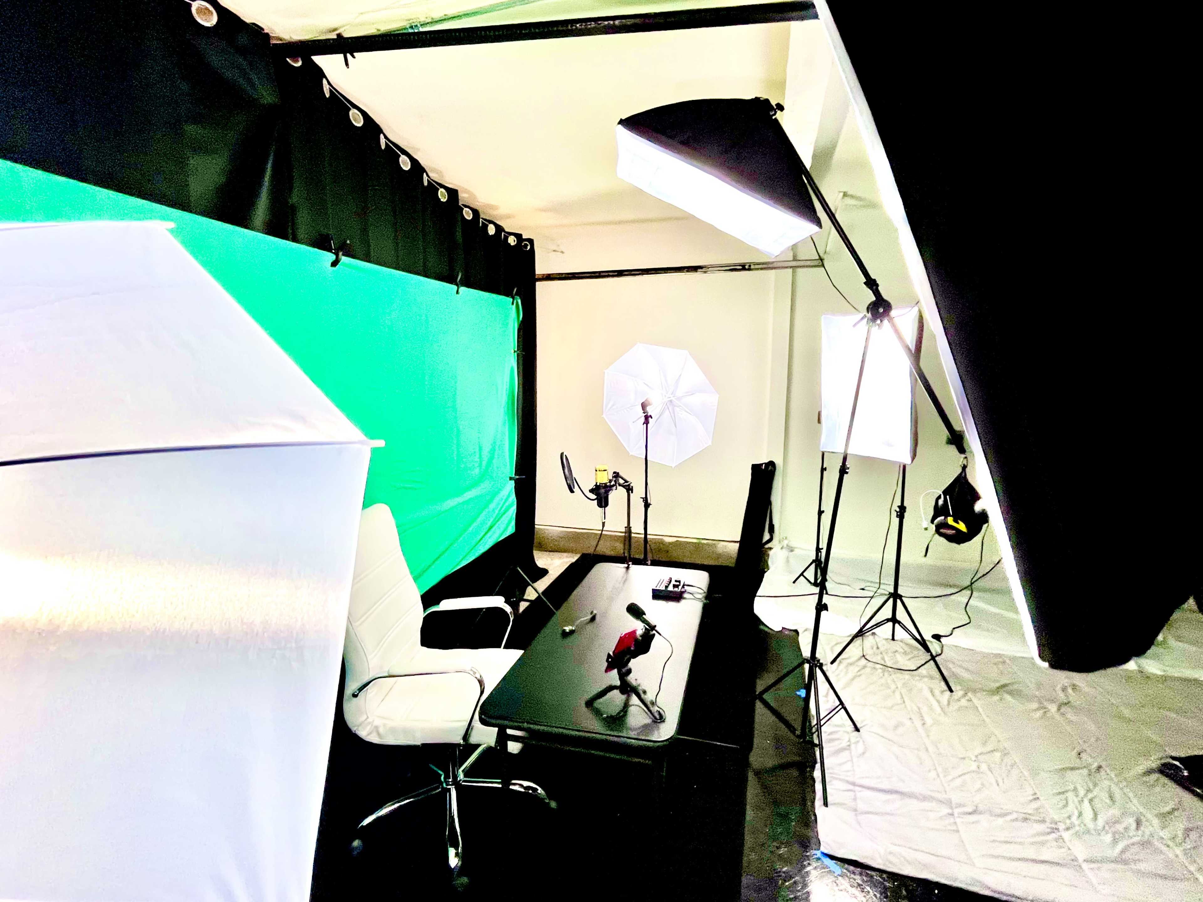 A video recording setup with a black desk, a white chair, and multiple lighting sources, including softboxes, against a green backdrop.