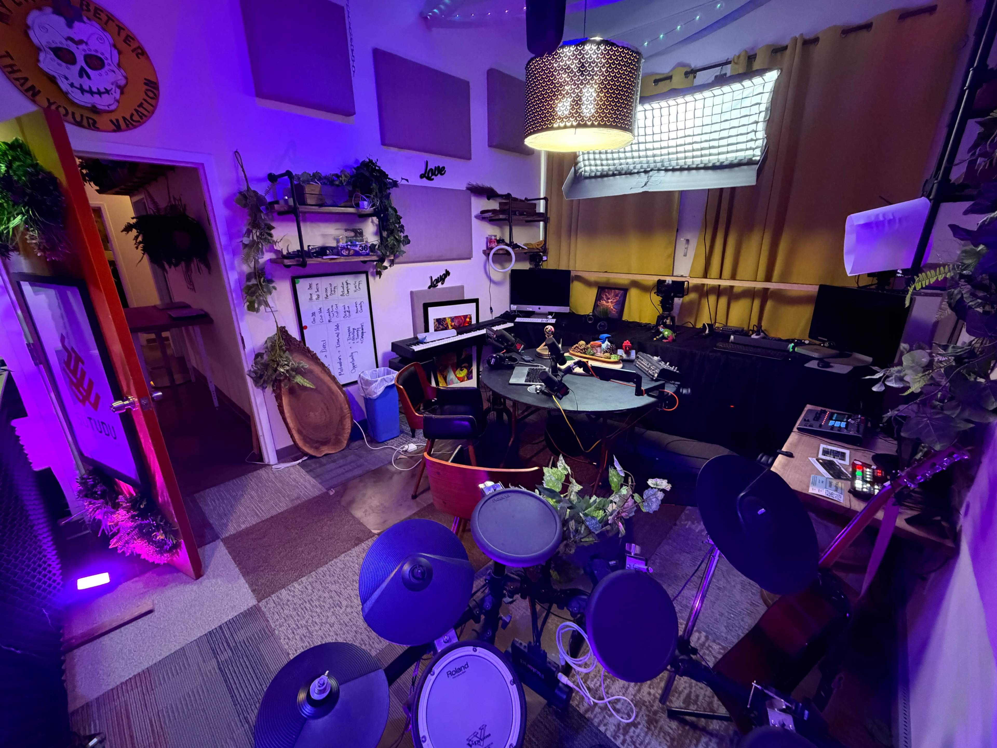 The image shows a colorful and well-decorated music studio equipped with electronic drums, a desk with various music equipment, and shelves adorned with plants and decor.