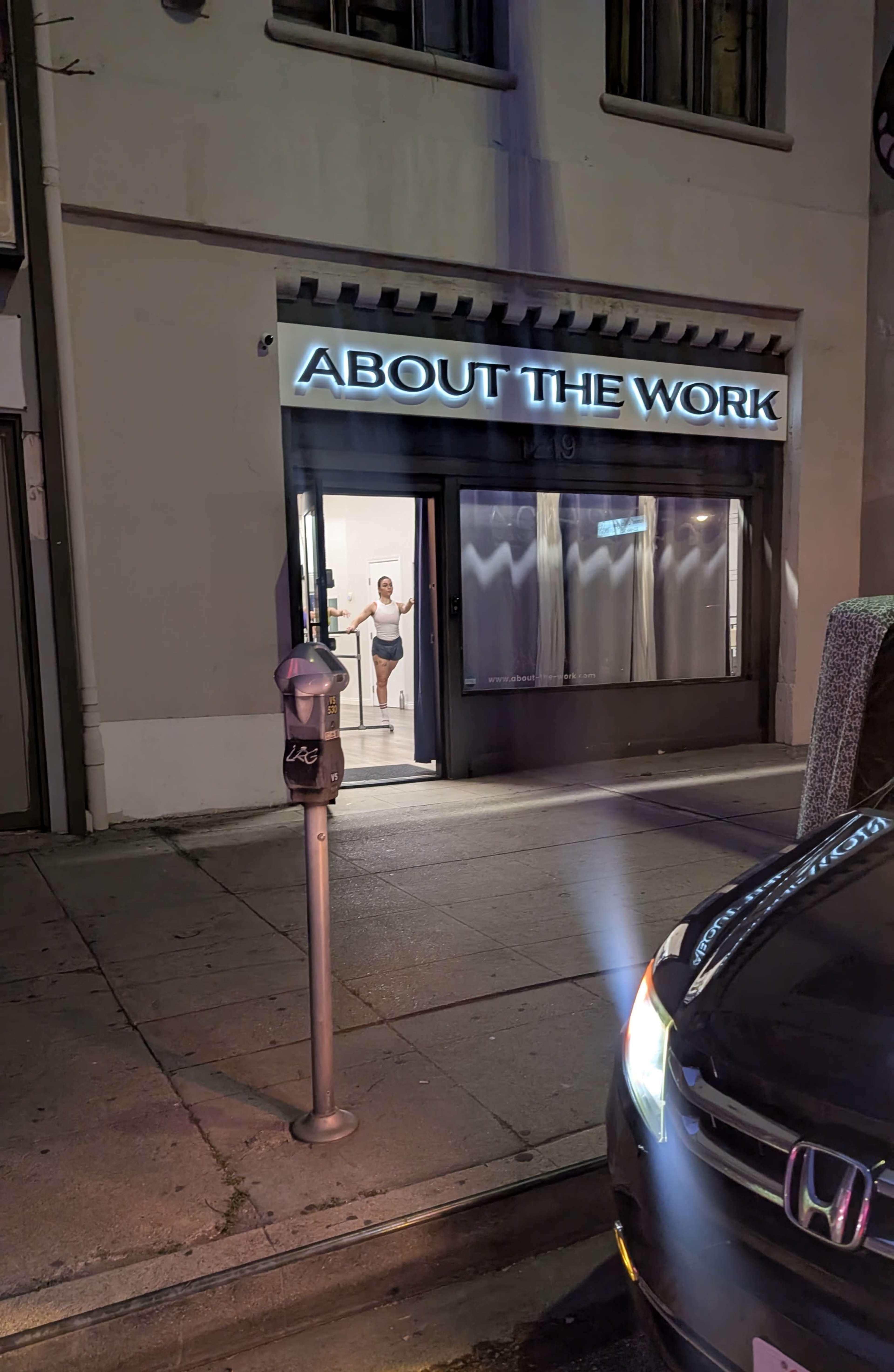 The image shows a storefront labeled "ABOUT THE WORK," with a person visible through the window and a parking meter nearby.