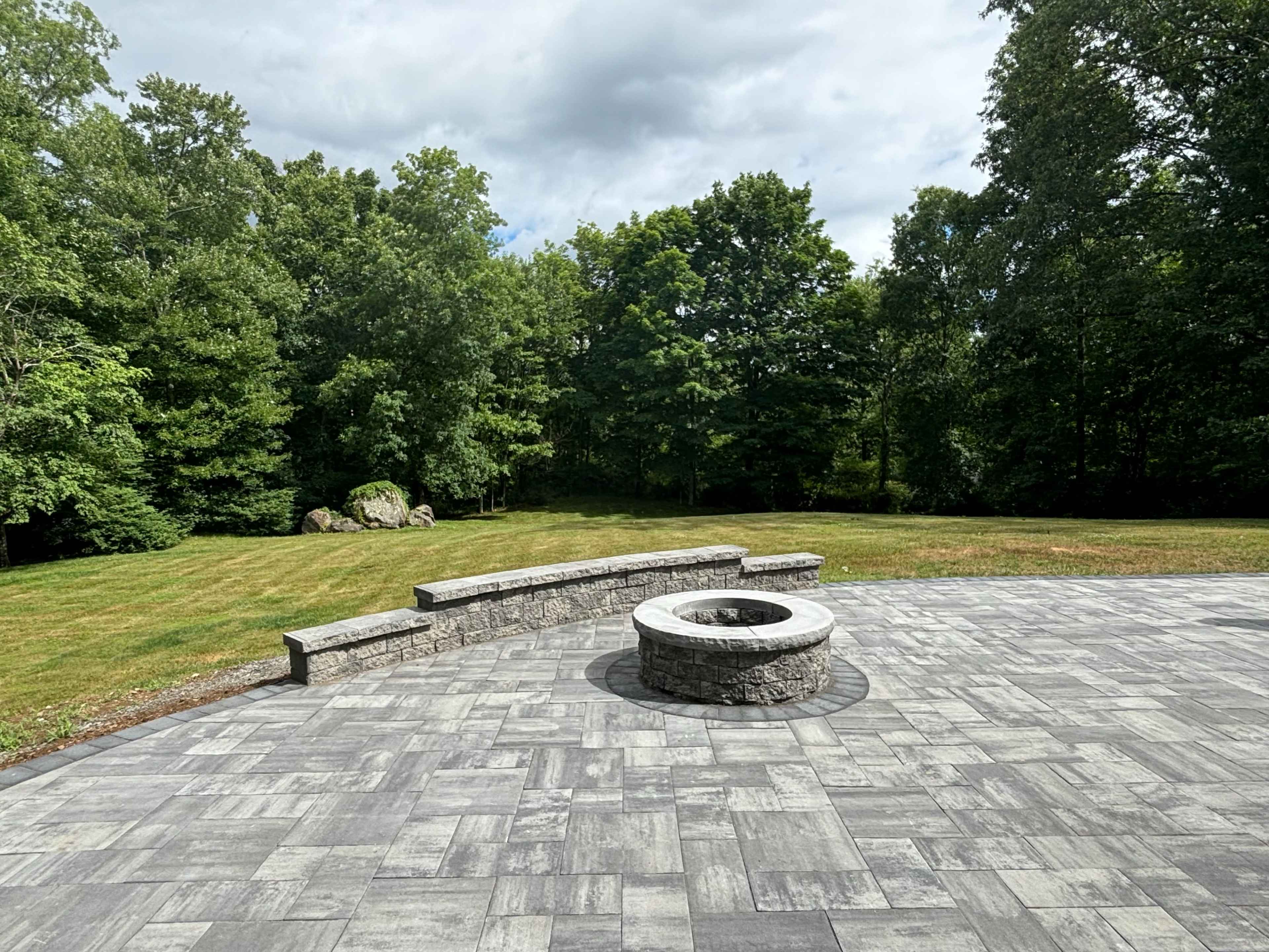 A stone fire pit is surrounded by a paved patio, with a grassy area and trees in the background.