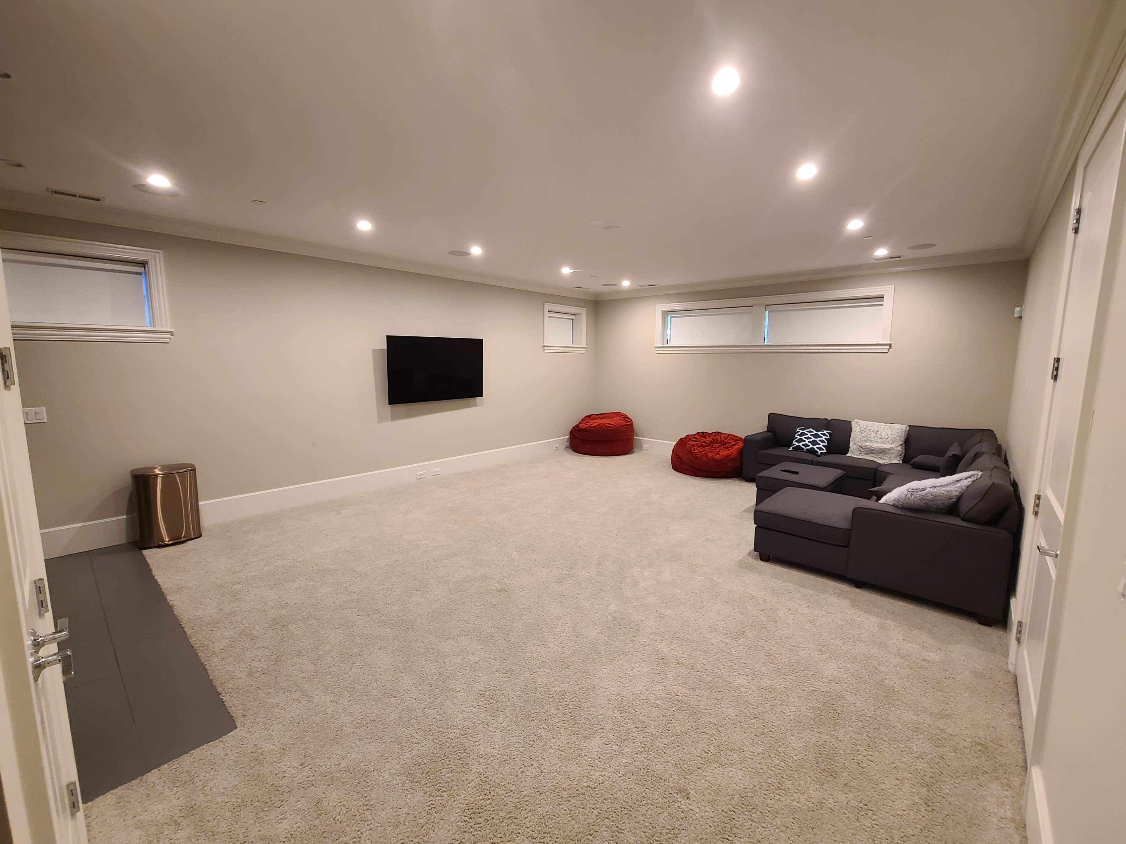 A spacious basement with beige carpet, a sectional couch, red bean bags, a mounted TV, and recessed lighting.
