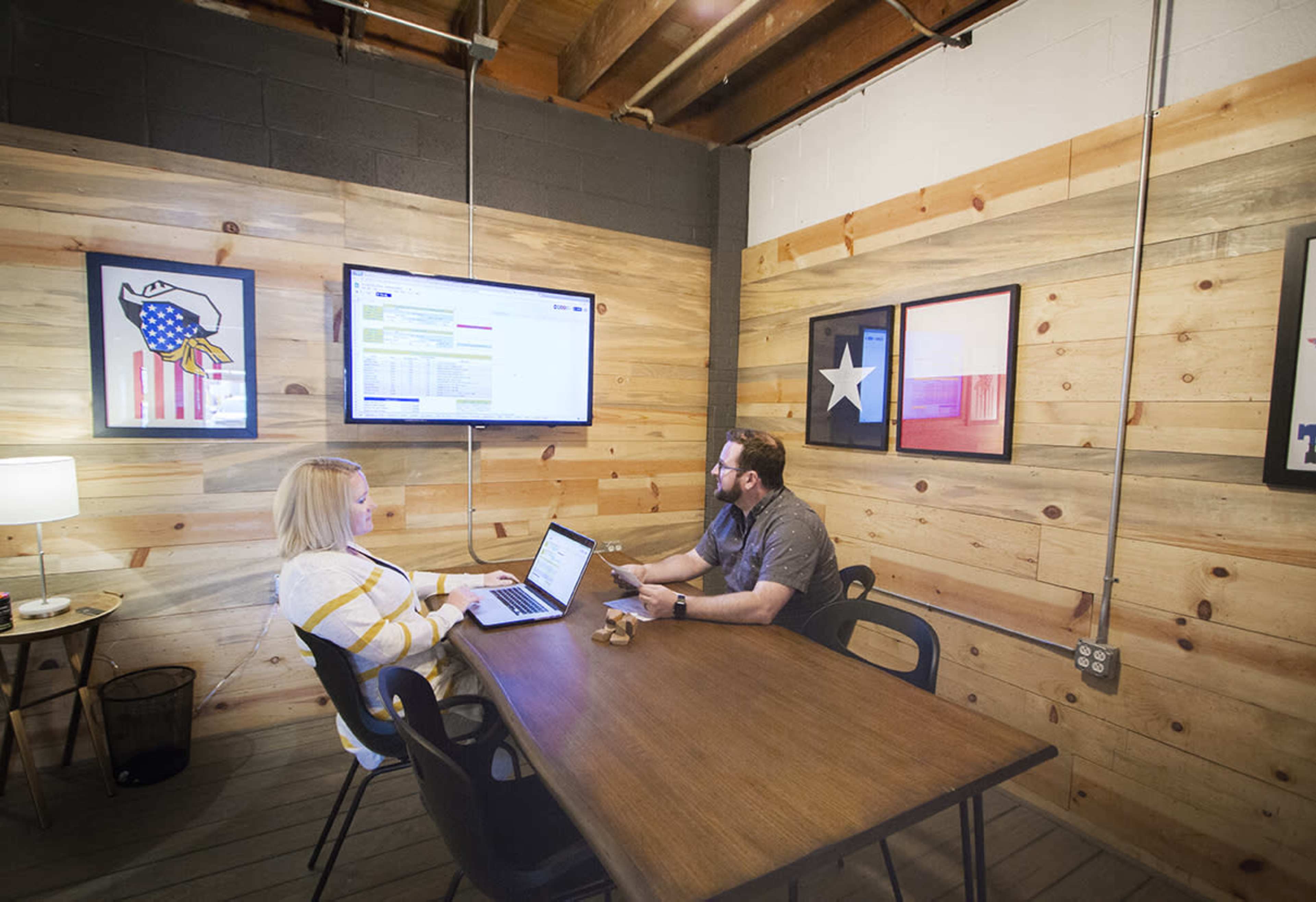 Two people are seated at a wooden table in a room with wooden walls, focusing on a laptop and a monitor displaying information.