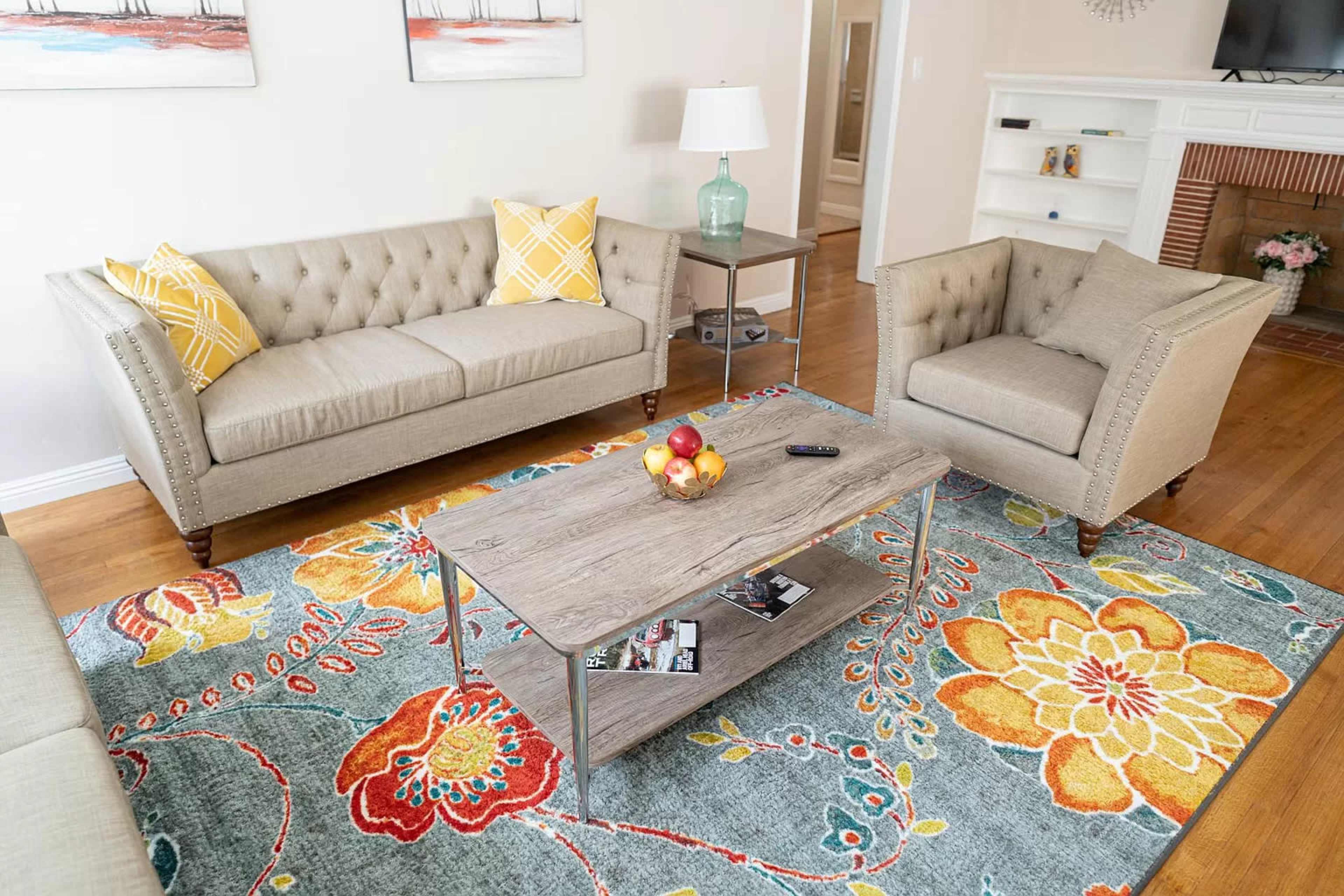 The image shows a living room with a beige tufted sofa, an armchair, a wooden coffee table, and a colorful area rug featuring floral patterns.