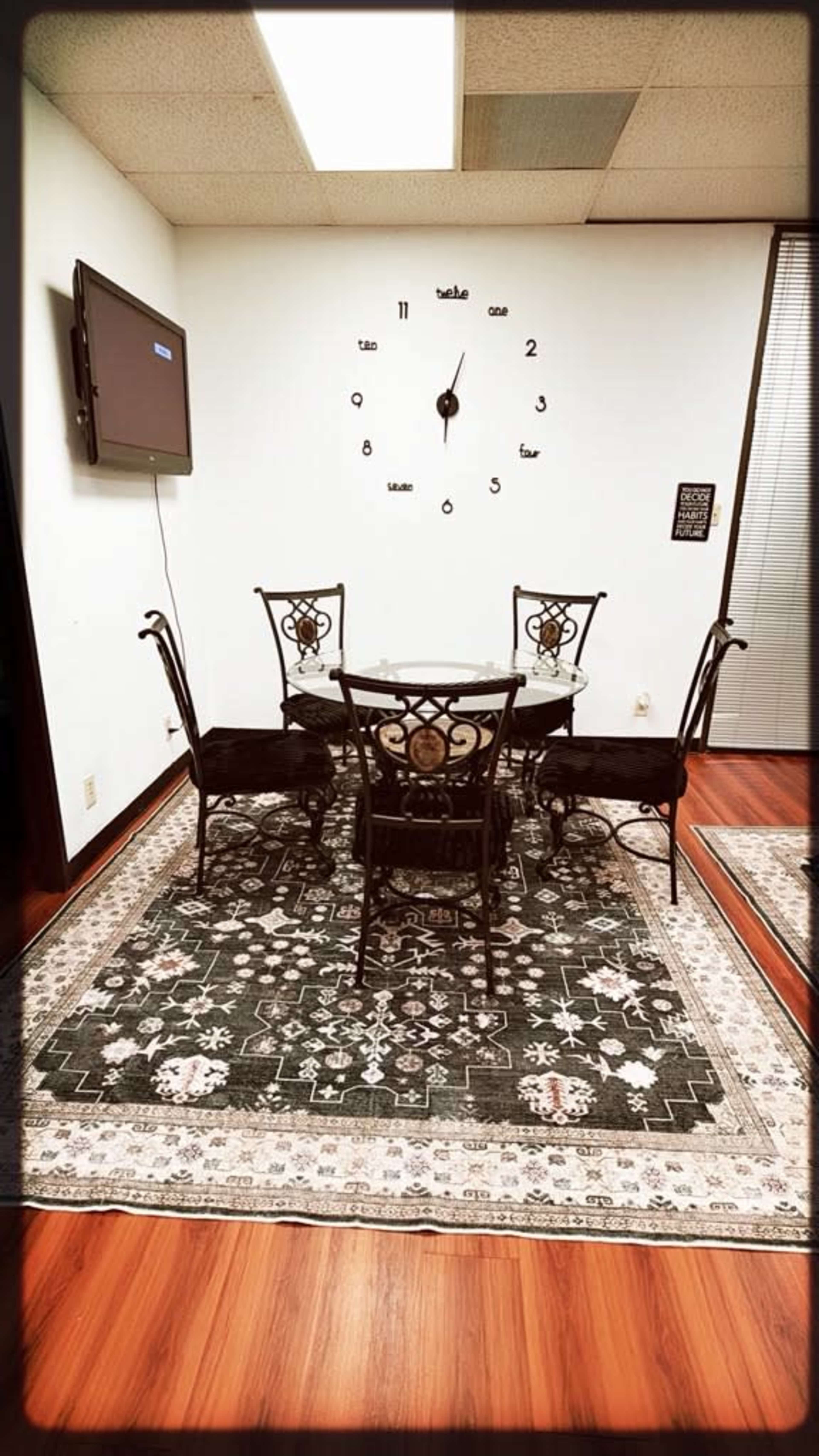 The image shows a small dining area with a round table surrounded by four wrought iron chairs, a large wall clock, and a television mounted on the wall.