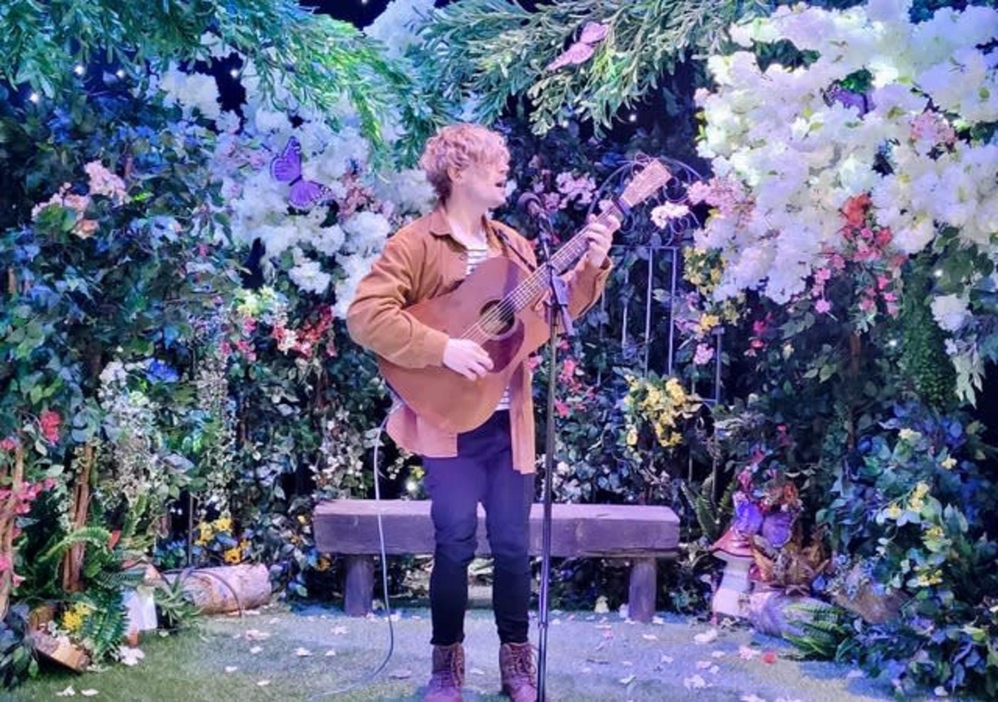 A musician plays an acoustic guitar on a stage surrounded by lush floral decor and greenery.