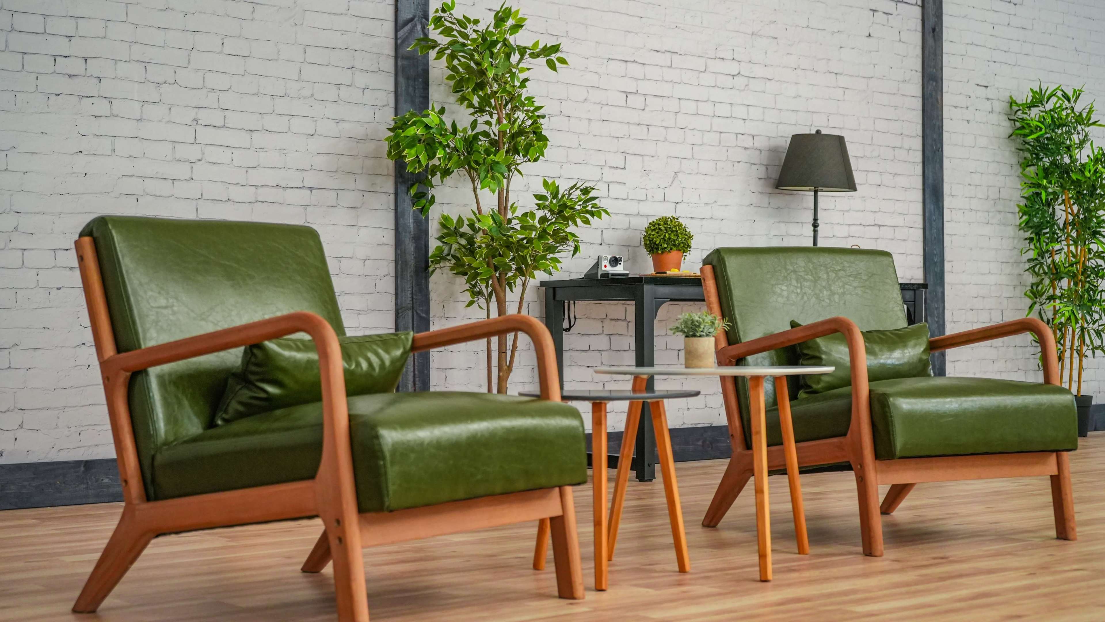 Two green armchairs with wooden frames positioned around a small round table in a room with a light brick wall and potted plants.