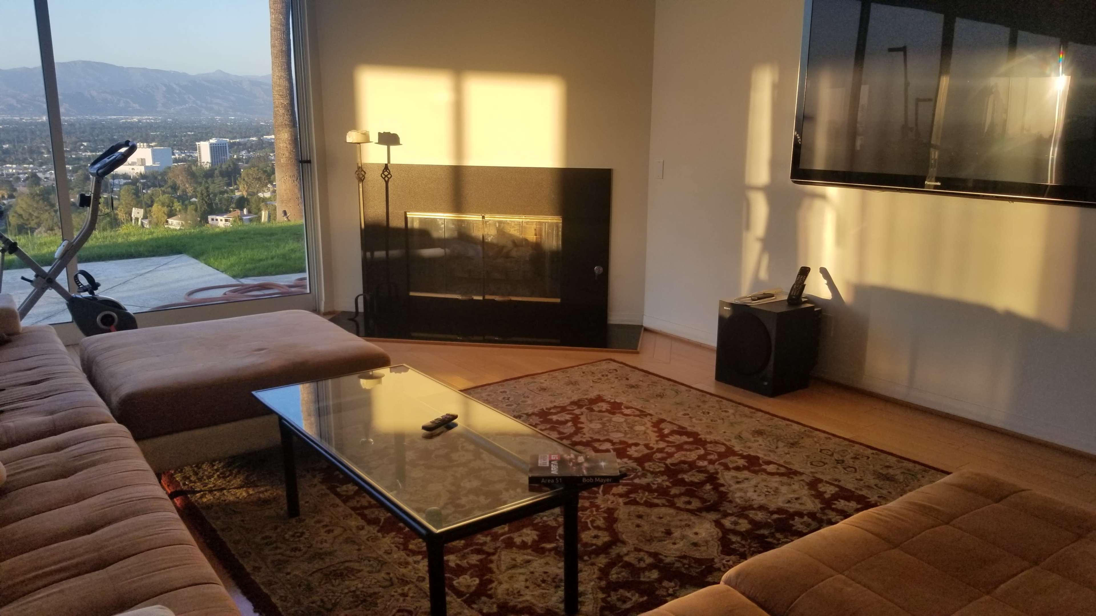 A living room with a sofa, glass coffee table, and a wall-mounted television, filled with sunlight and featuring a view of distant mountains beyond a window.